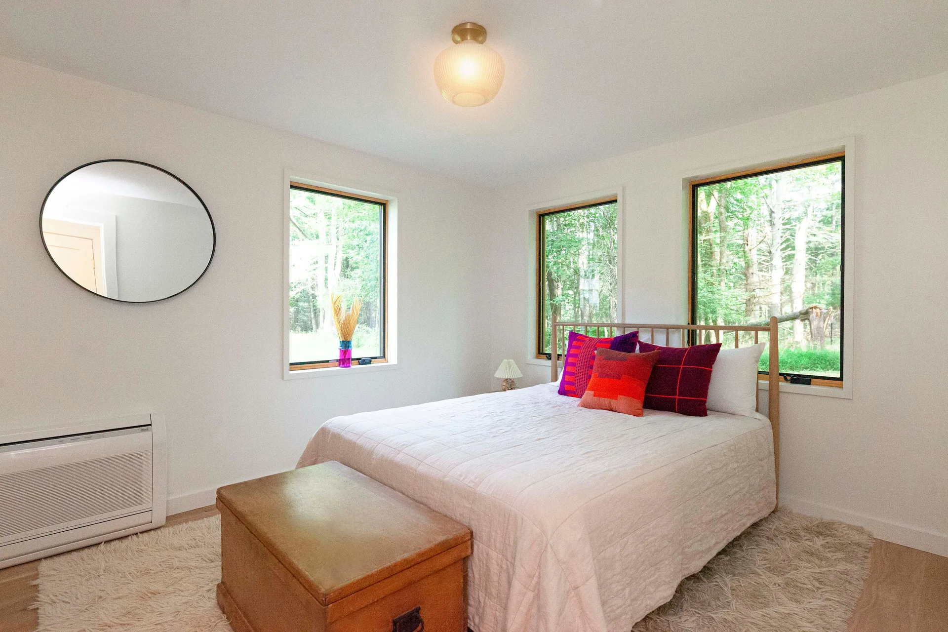 A bright white bedroom features a wooden bed frame with colorful pillows, a shag rug, a wooden chest, and two windows showing forest views.