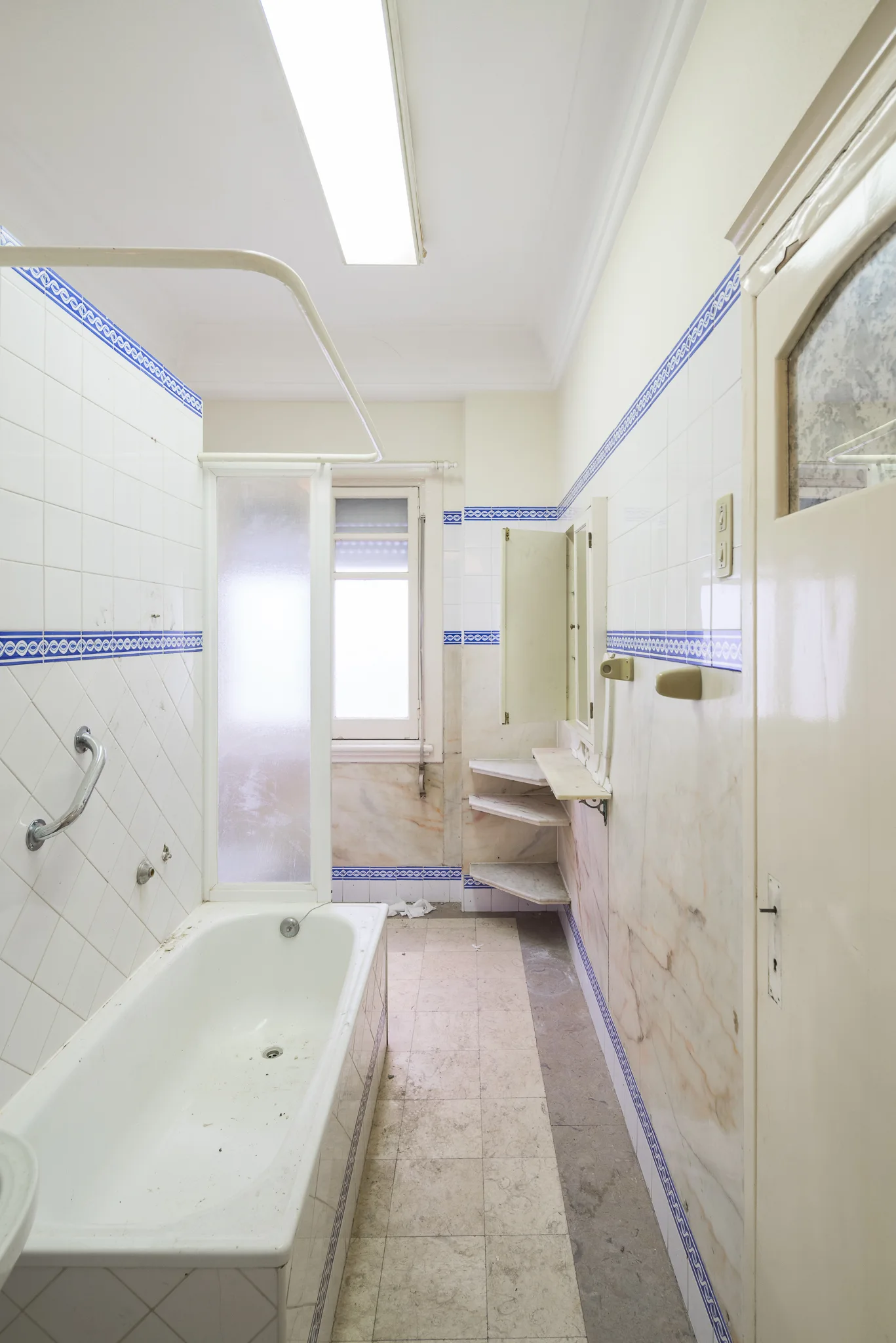 A narrow vintage bathroom features a bathtub, white wall tiles with a blue patterned border, and marble-look lower wall panels.