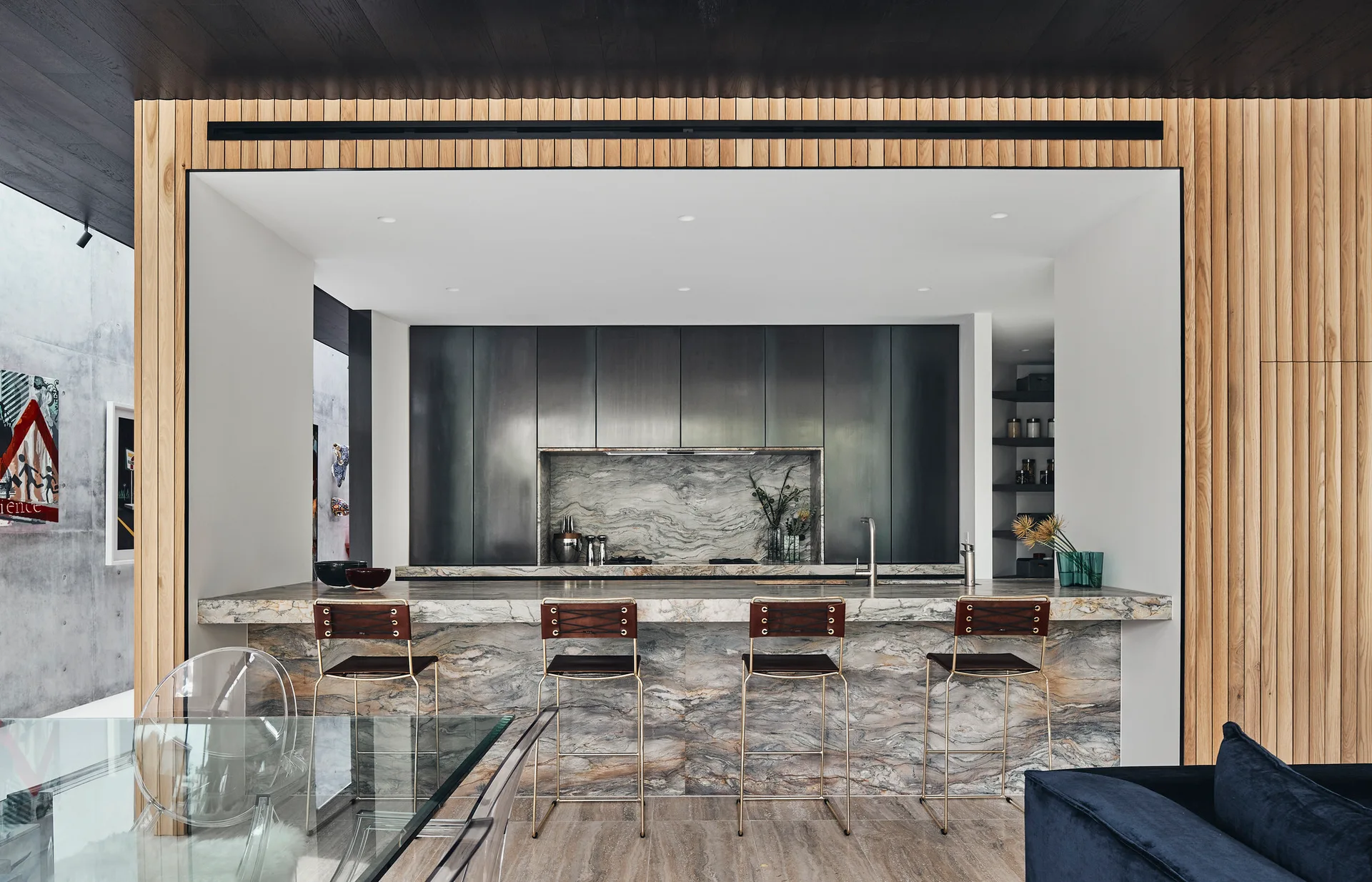 A modern kitchen features a marble-clad island with leather bar stools set against a backdrop of dark cabinetry and vertical wood slat paneling.