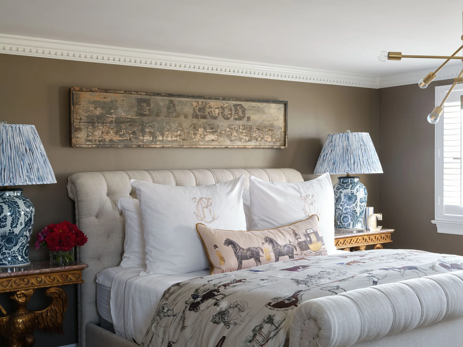 A bedroom features a tufted headboard against a warm brown wall, decorated with a vintage wooden carriage sign and blue patterned lamps.