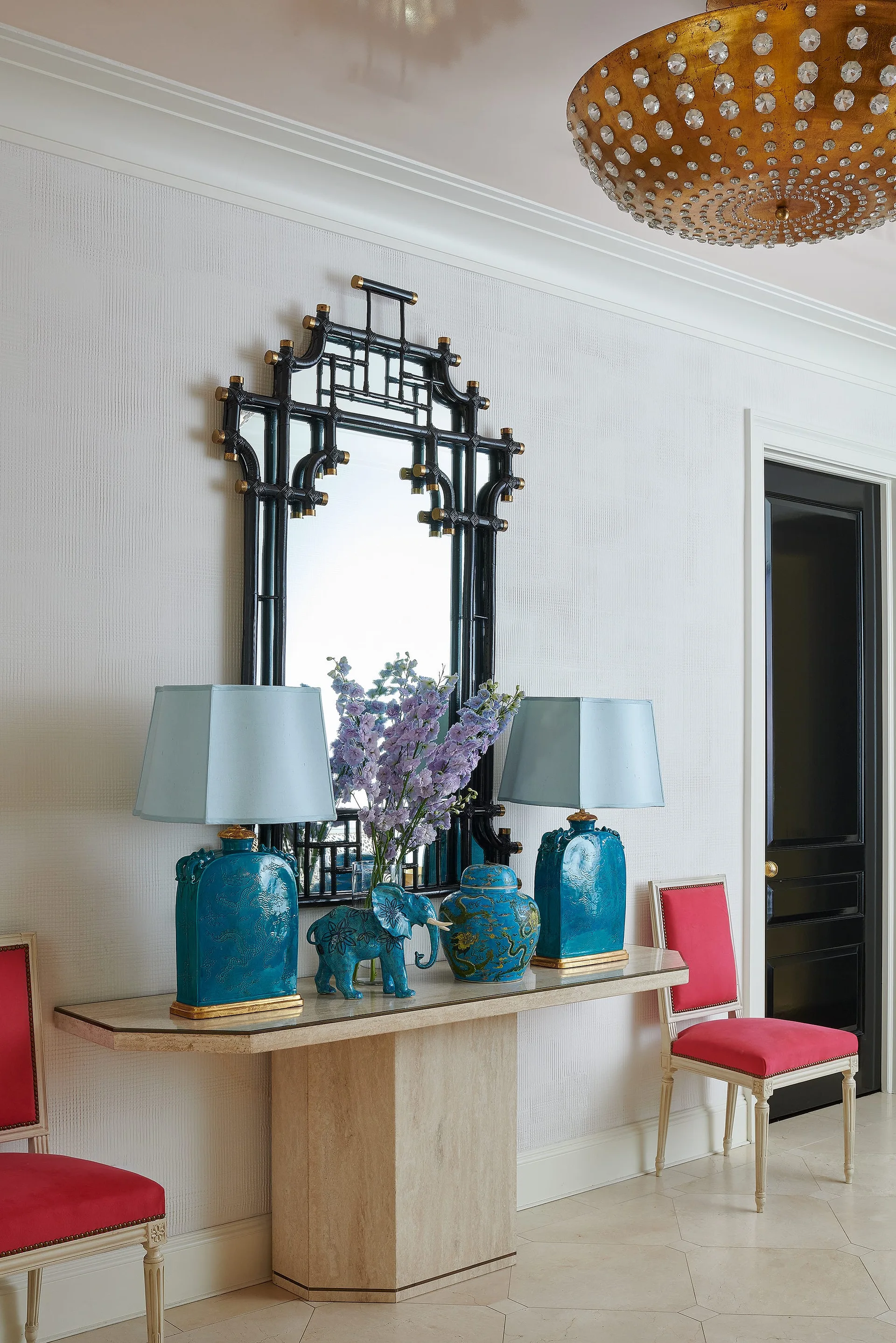 An entryway features a stone console table with two blue lamps and a black chinoiserie mirror, flanked by two pink upholstered chairs.