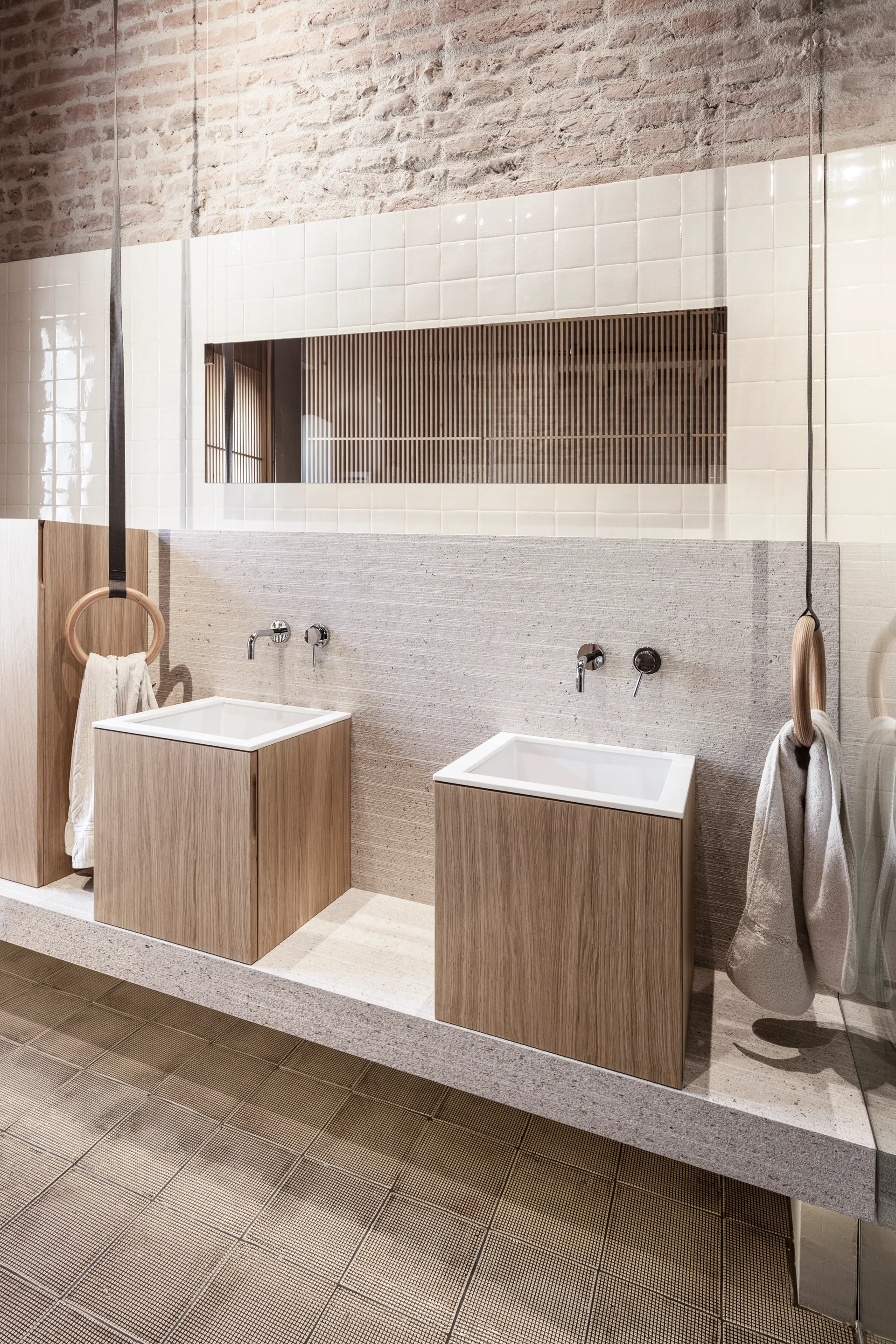 Two square wood block vanities sit on a long grey stone ledge beneath a cream tiled wall and exposed brickwork in a modern bathroom.