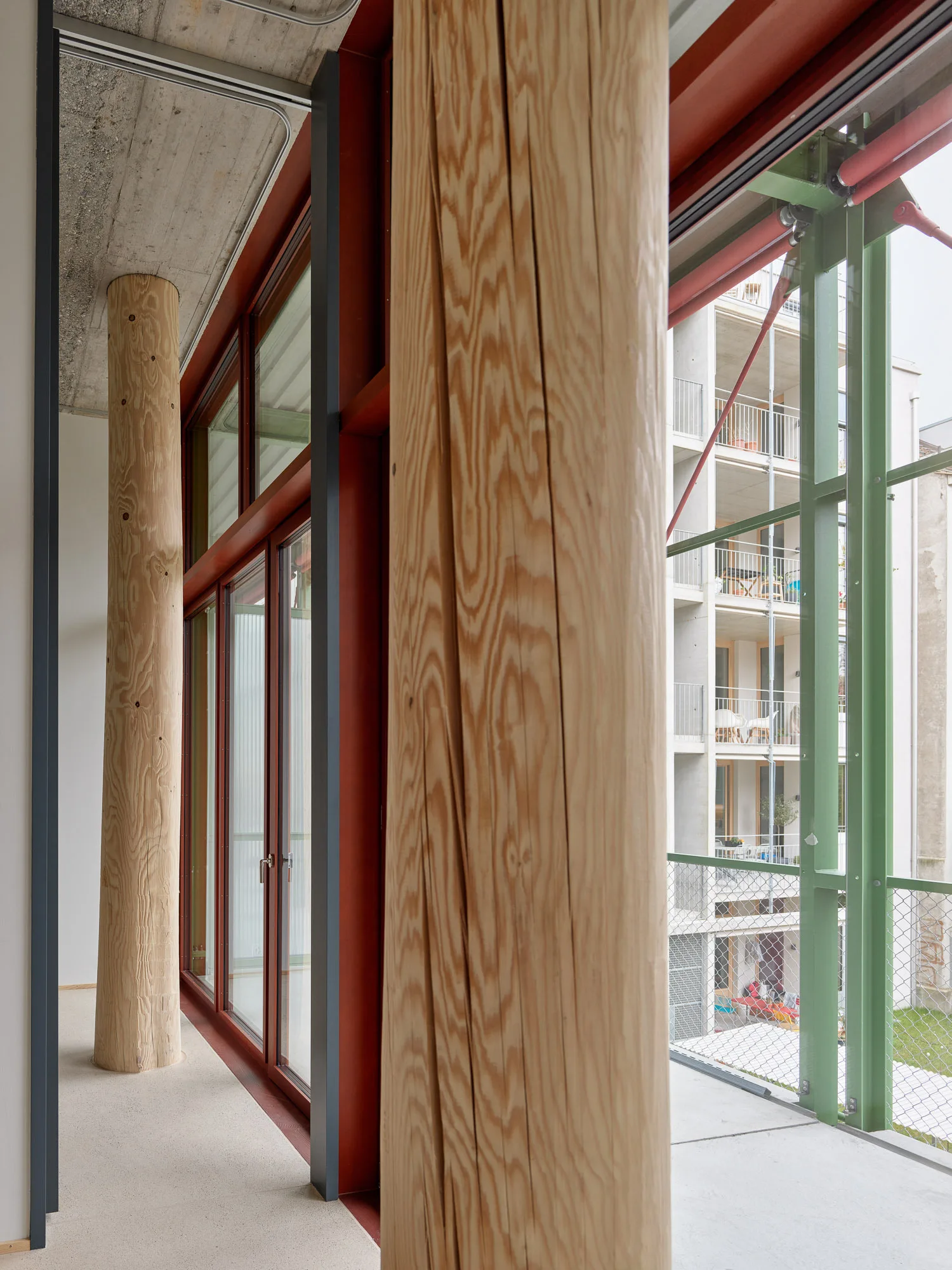 Raw timber columns stand next to large red-framed glass windows beneath an exposed concrete ceiling in an industrial interior.