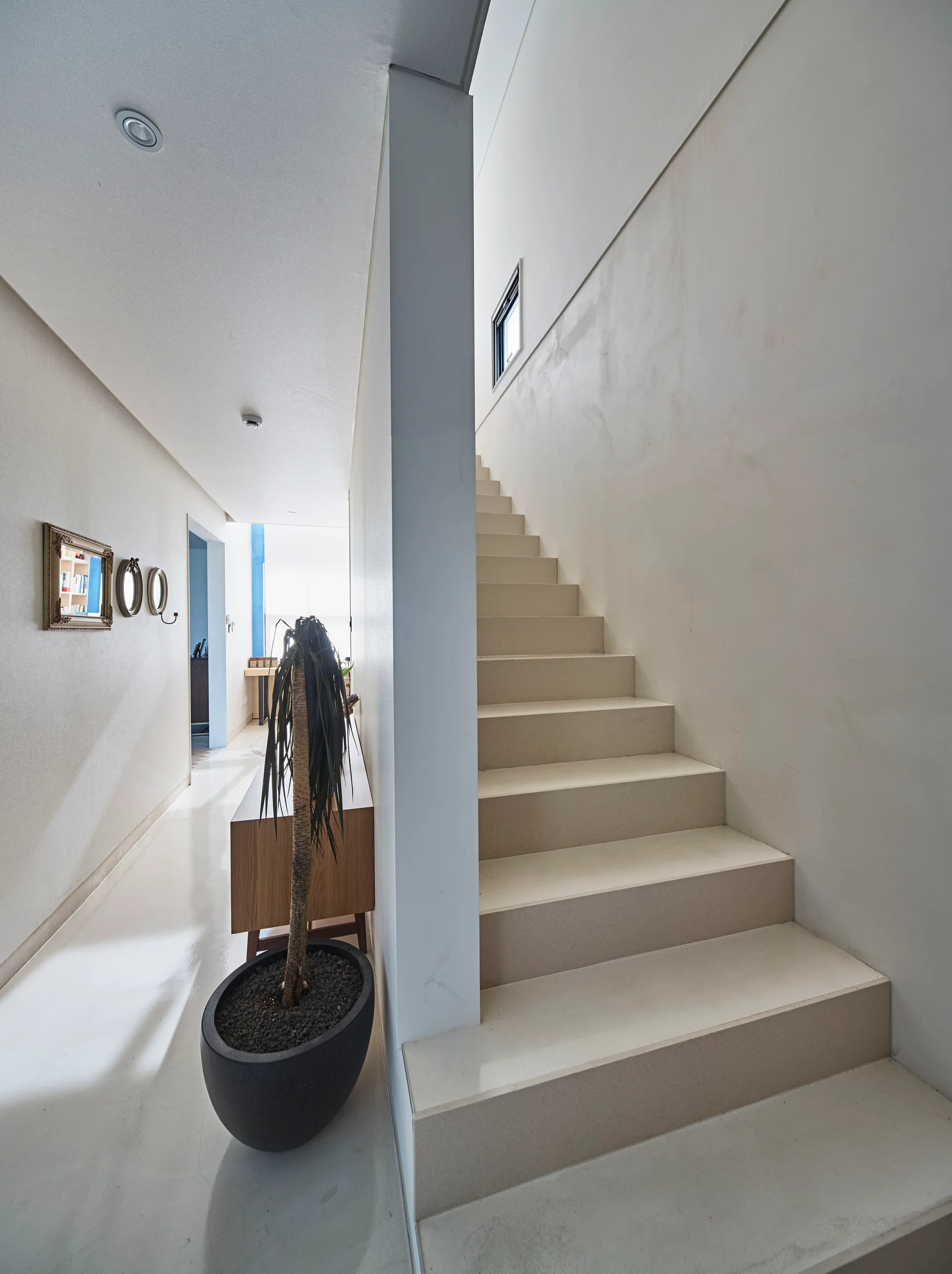 An off-white minimalist staircase stands next to a glossy white hallway featuring a tall potted plant and a wood sideboard.