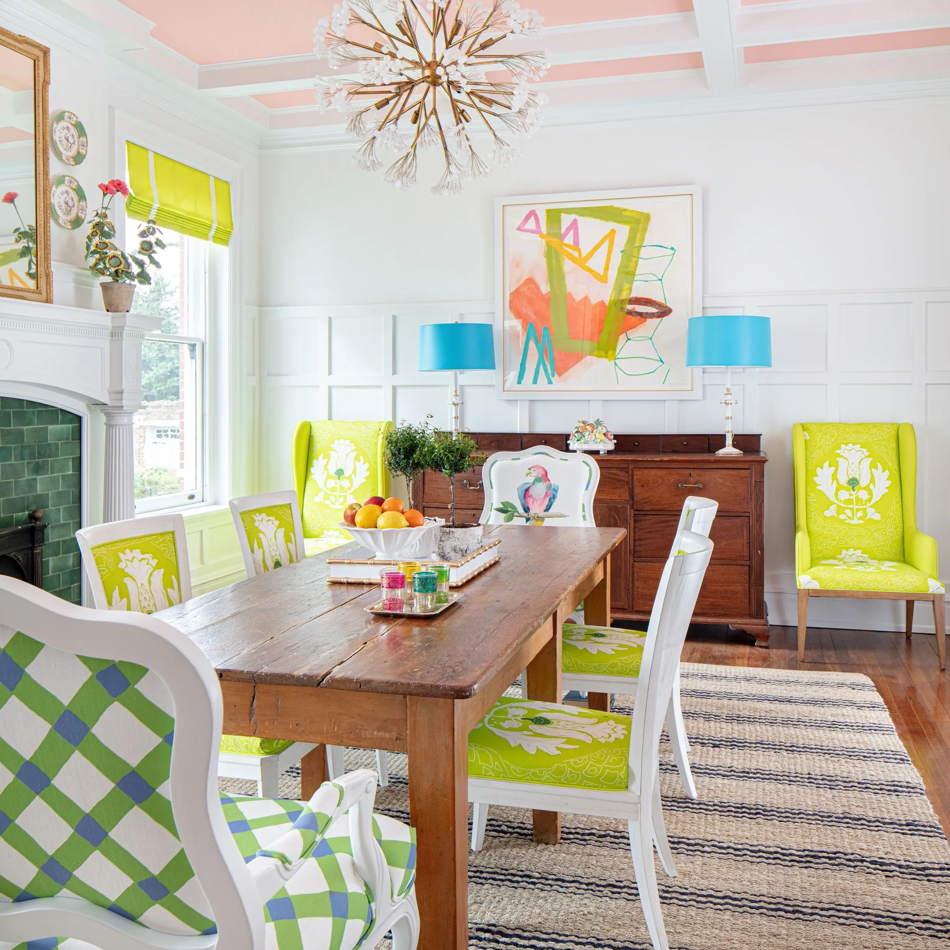 An eclectic dining room with a rustic wooden table, chairs upholstered in lime green patterns, white paneled walls, and a pale pink ceiling.
