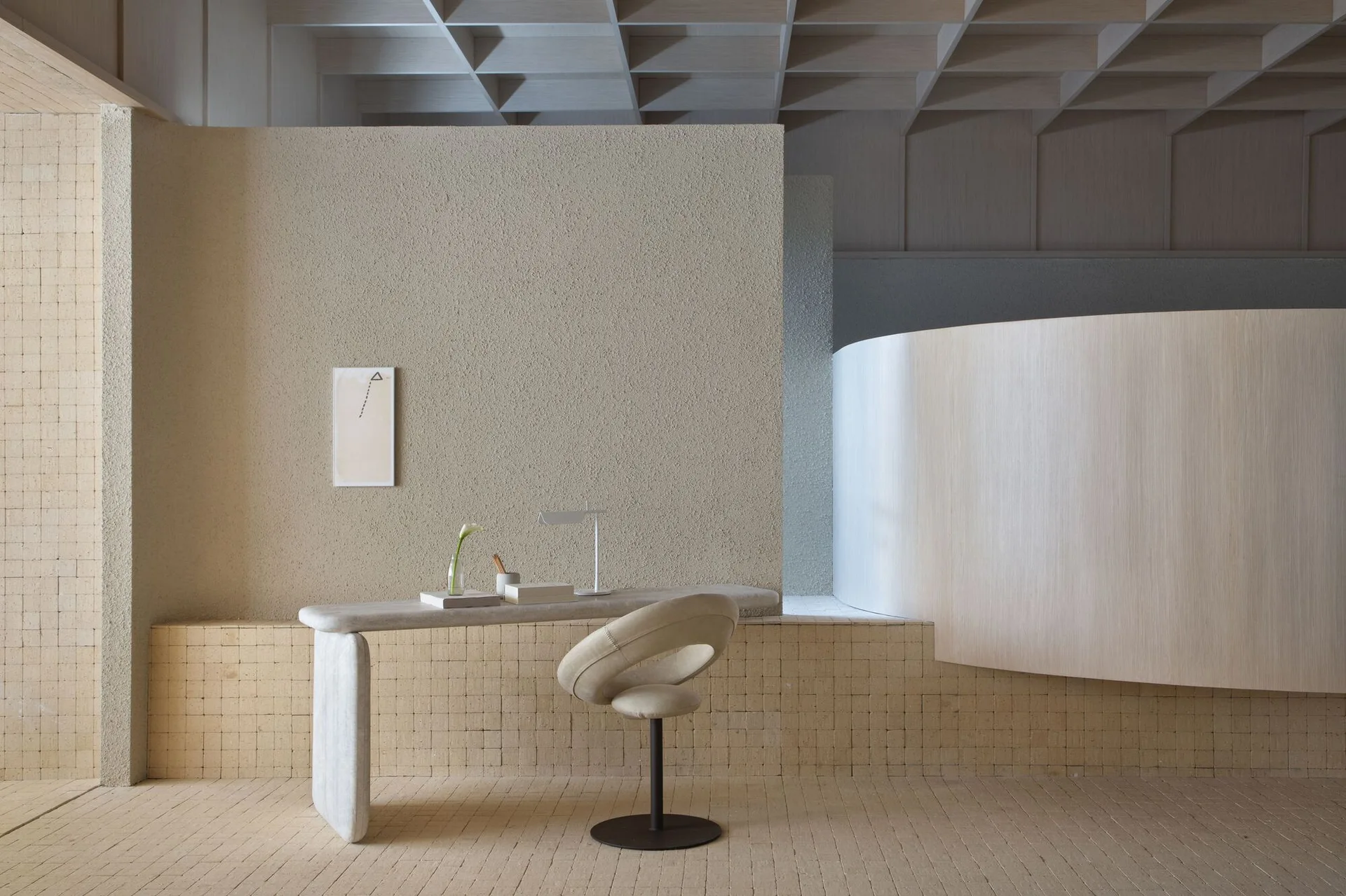 A minimalist home office features a sculptural stone desk, an ivory swivel chair, and textured beige walls under a grid coffered ceiling.