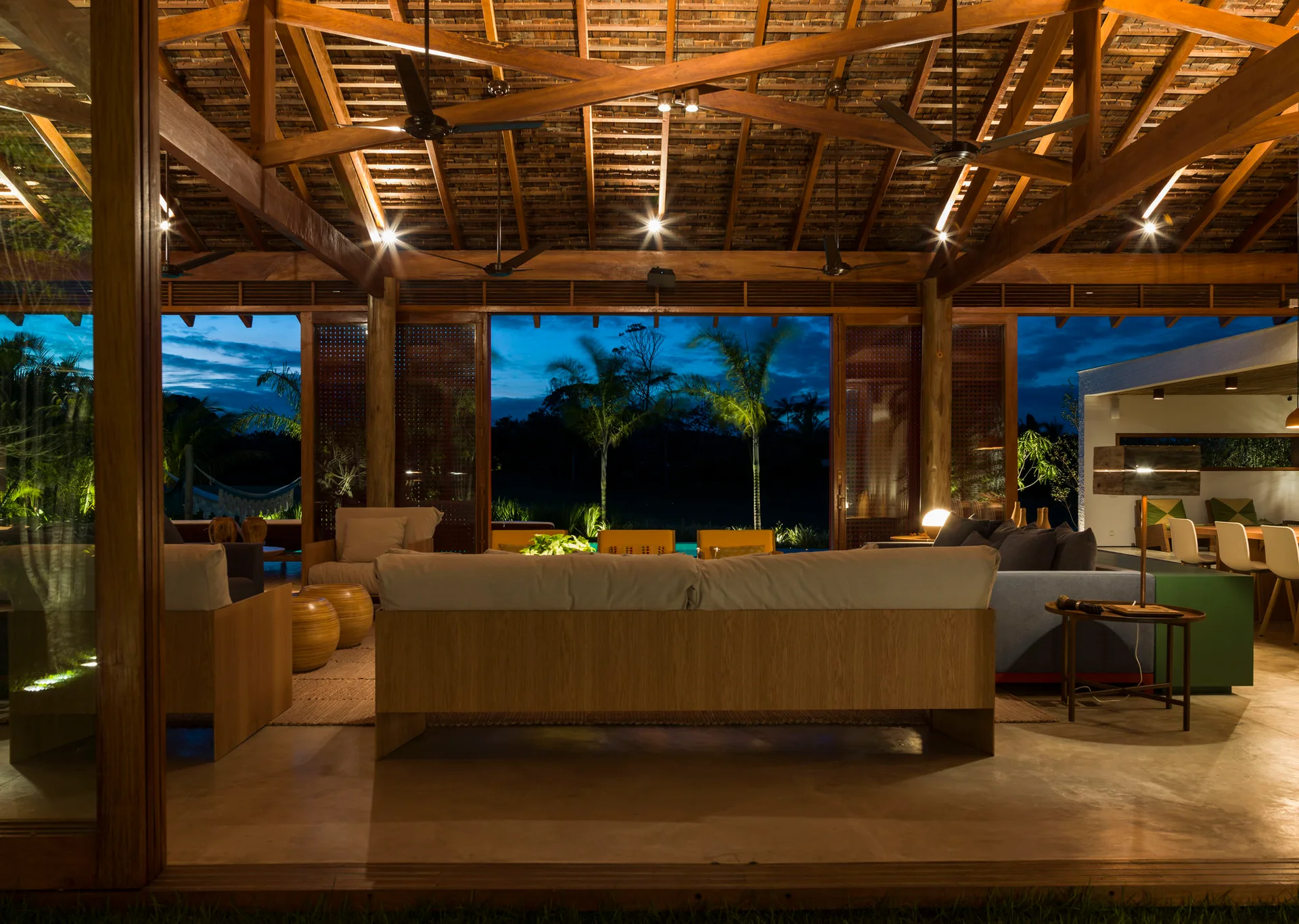 An open-air pavilion lounge features low-profile sofas under a high, vaulted wooden beam ceiling illuminated by warm spotlights at dusk.