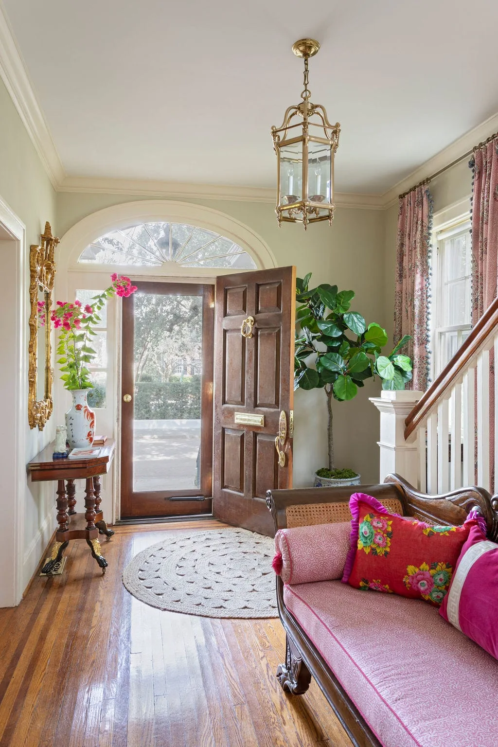 A traditional entryway features pale green walls, a pink upholstered bench, a round woven rug, and a classic brass lantern hanging near the staircase.