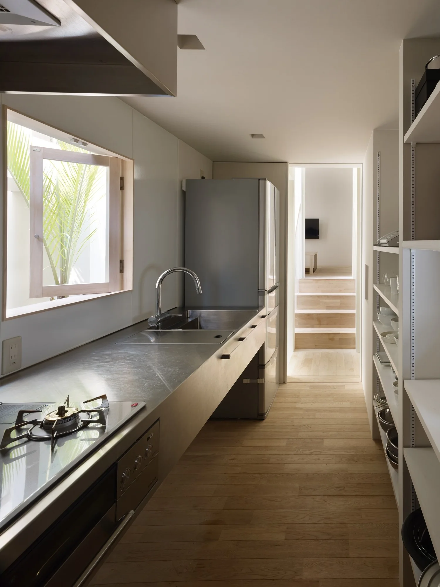 A minimalist galley kitchen with a long stainless steel countertop, a built-in cooktop, open white shelves, and a square window.