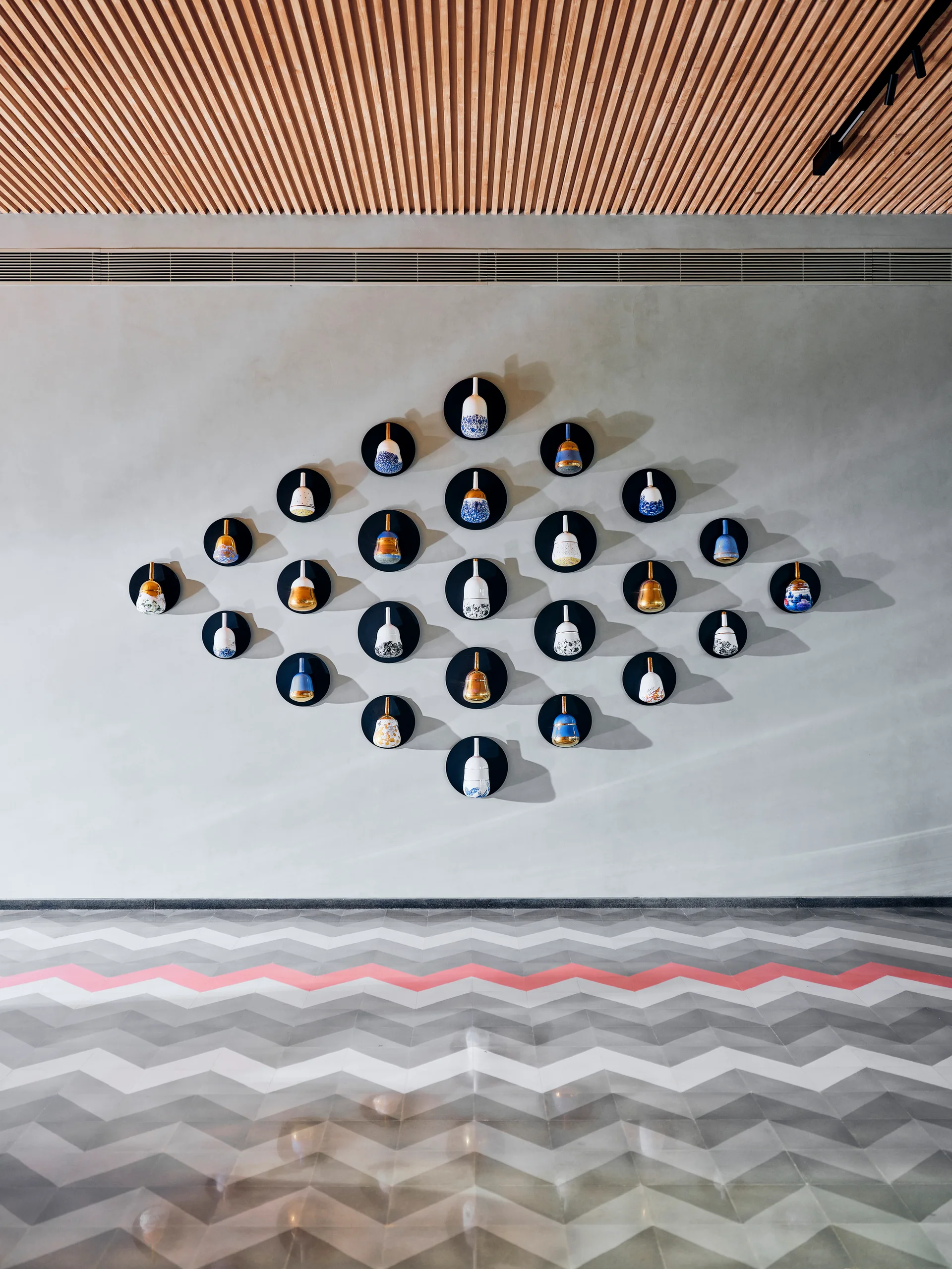 A grey accent wall features a diamond grid of small ceramic vessels on black mounts below a wood slatted ceiling and above a chevron tile floor.