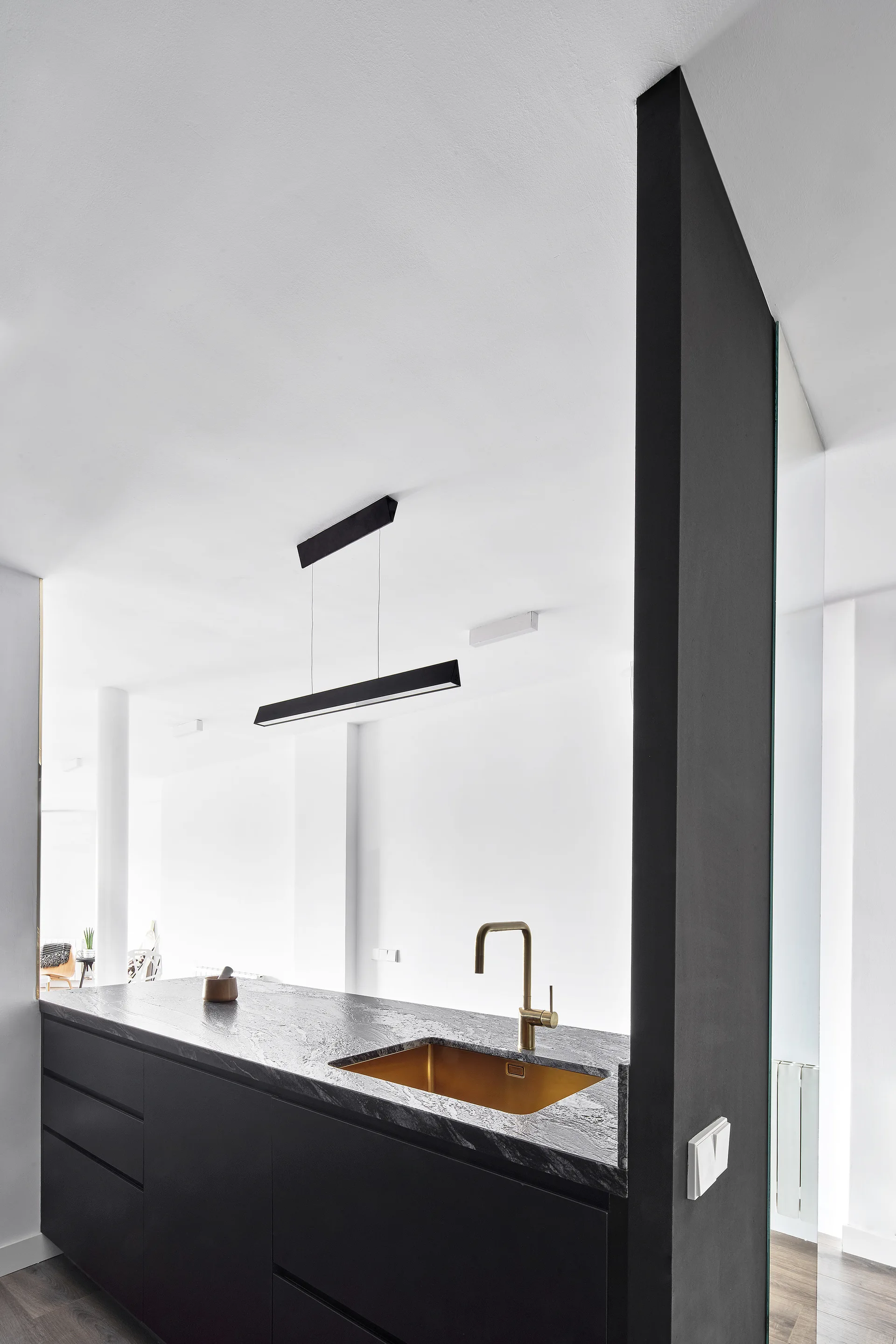 A minimalist black kitchen island with a dark stone countertop, integrated gold sink, brass faucet, and a black linear pendant light.