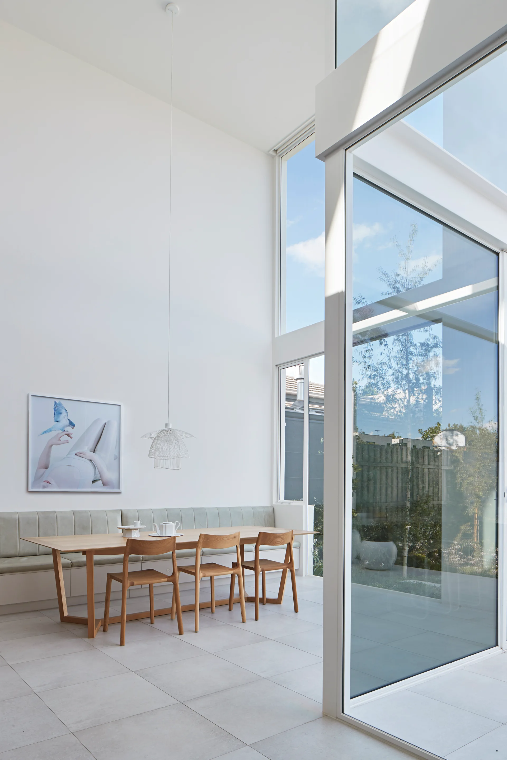 A bright minimalist dining room features a light oak table and chairs set against a long pale green upholstered banquette beneath a high ceiling.