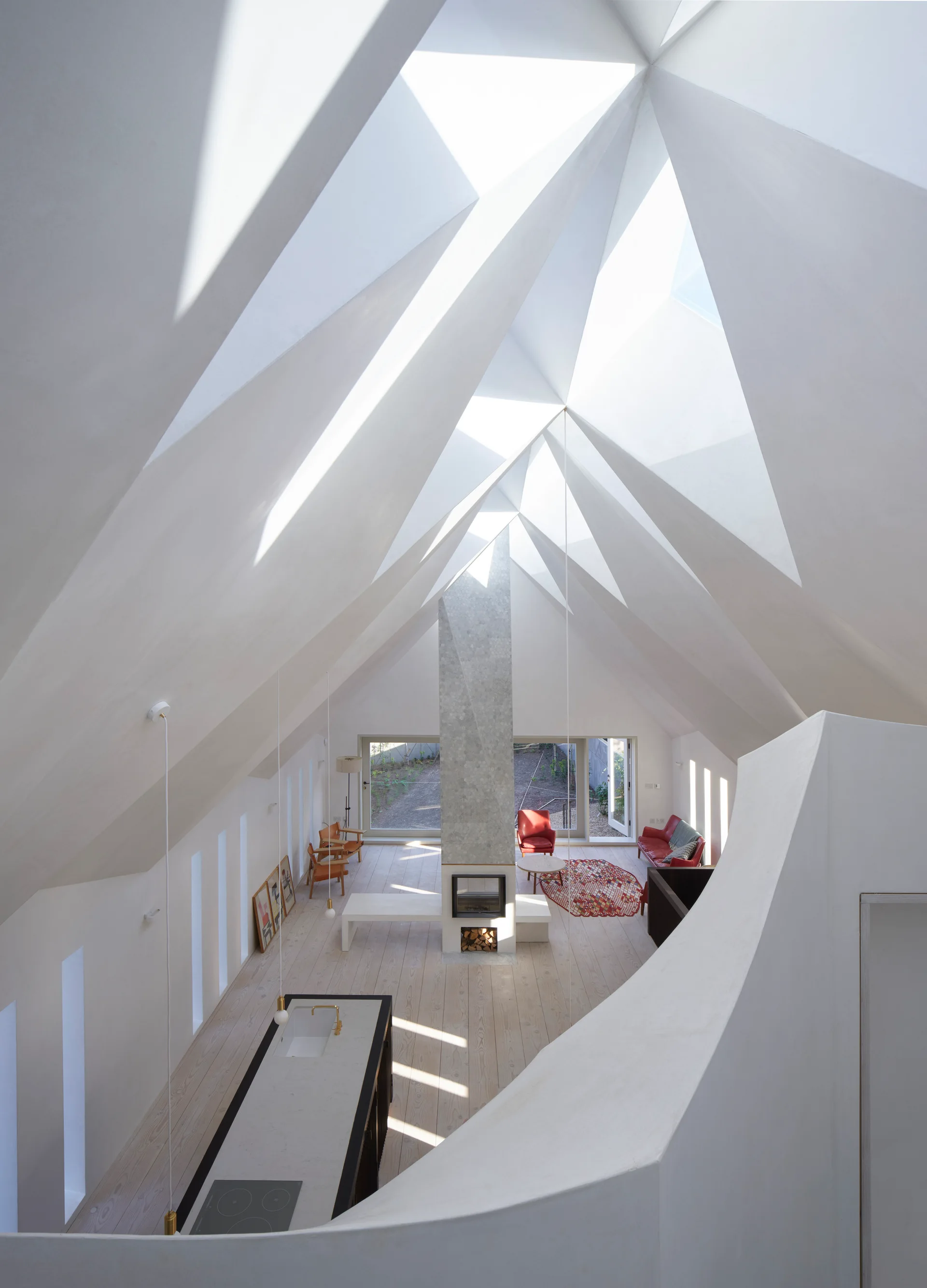 High-angle view of an open-plan living area featuring a white angular vaulted ceiling with skylights, a central stone fireplace, and pale wood floors.