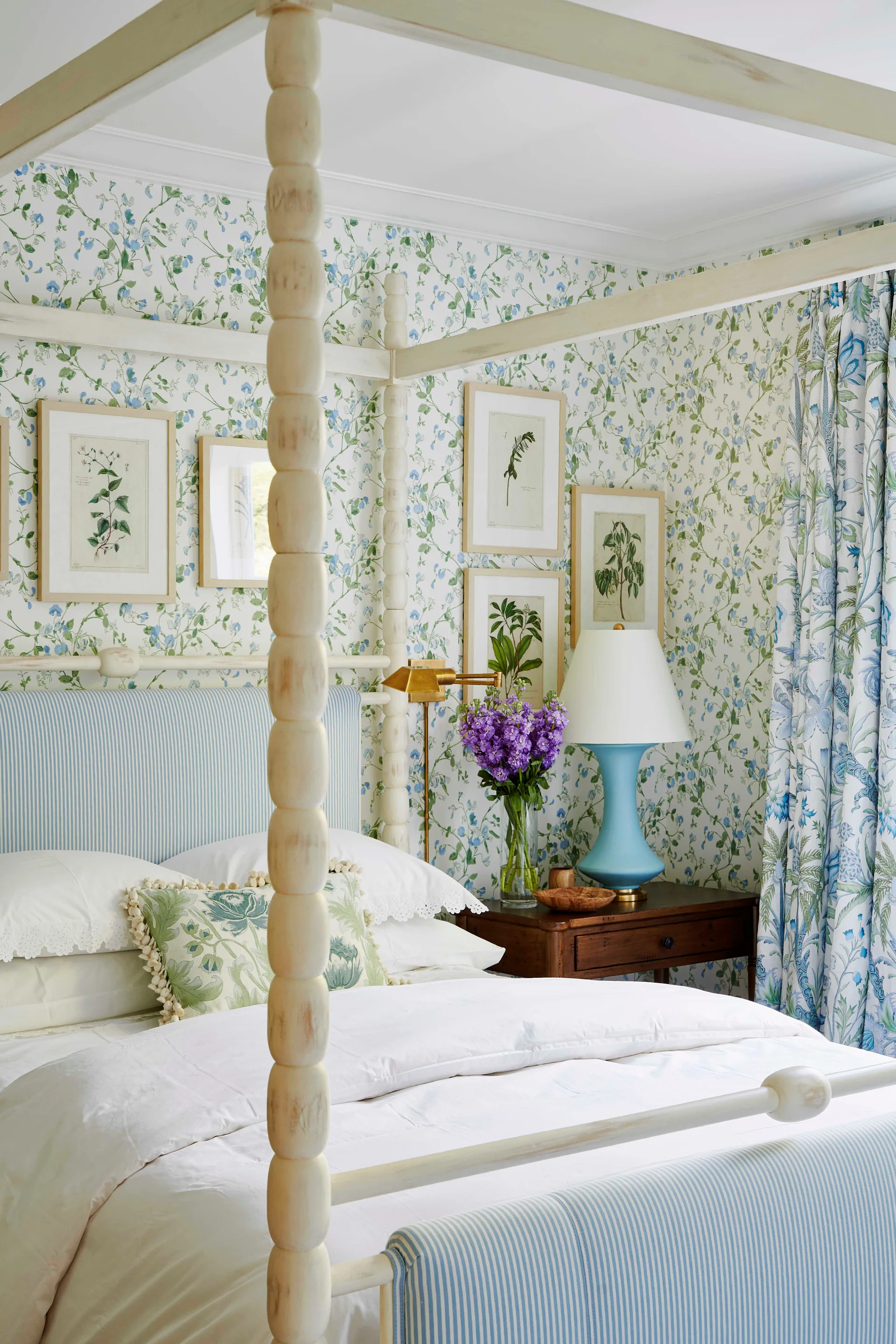 A traditional bedroom features a wooden spool four-poster bed against blue and green botanical wallpaper with framed botanical art.