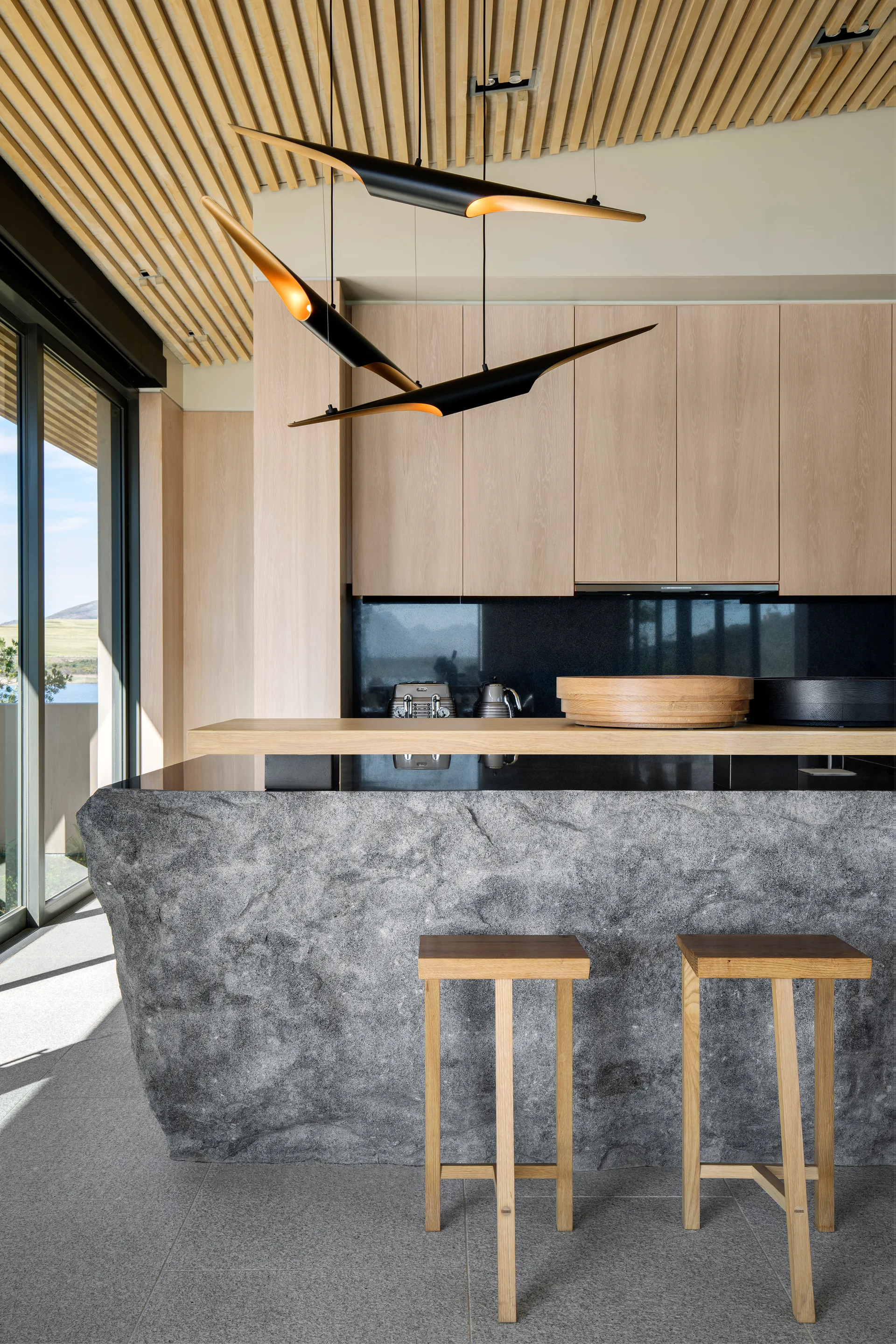 A modern kitchen featuring a textured grey stone island, wooden bar stools, light wood cabinets, and a slatted timber ceiling.