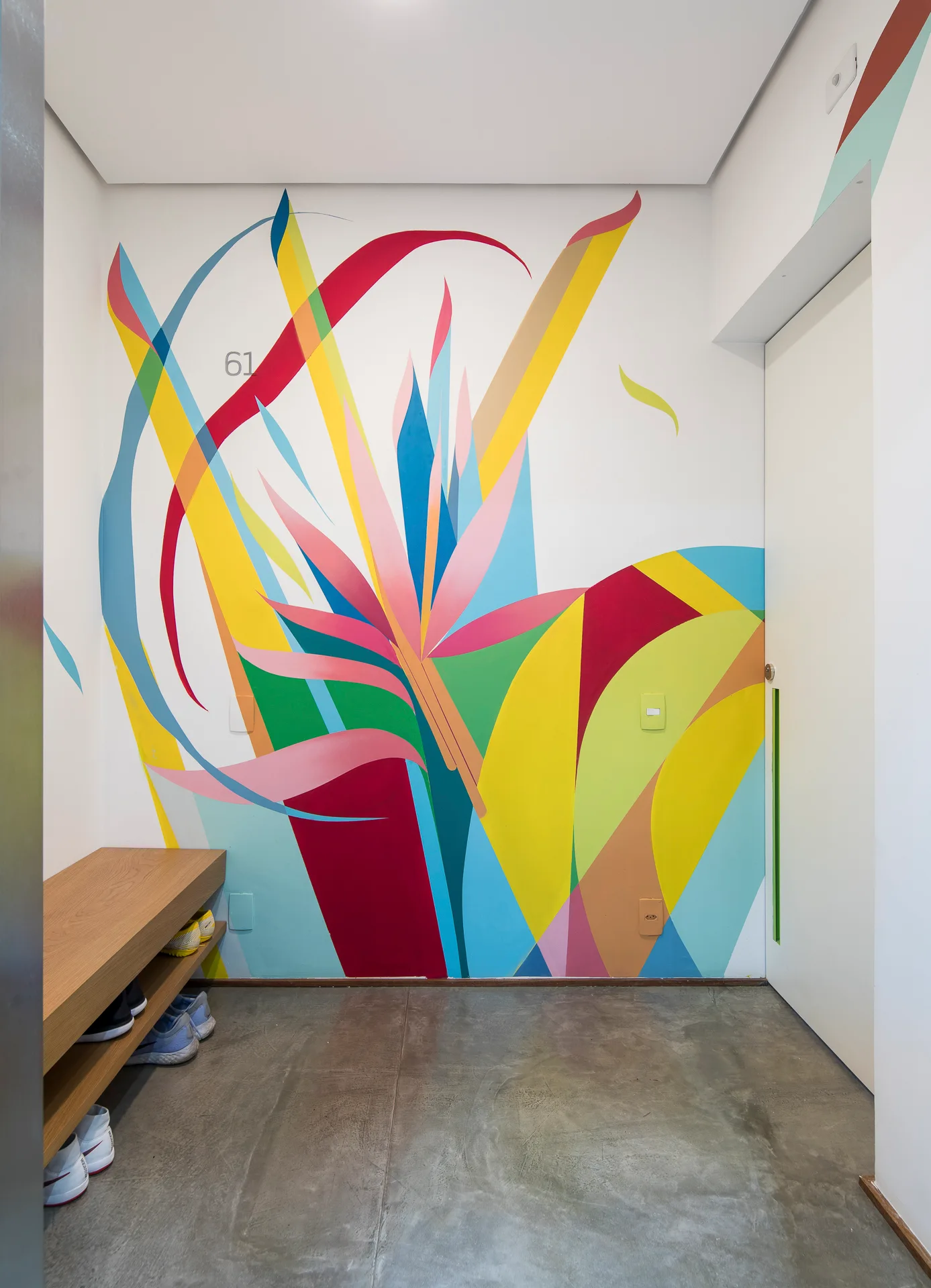 A modern entryway features a vibrant multicolored floral mural painted on a white wall next to a wooden shoe bench and grey concrete floor.
