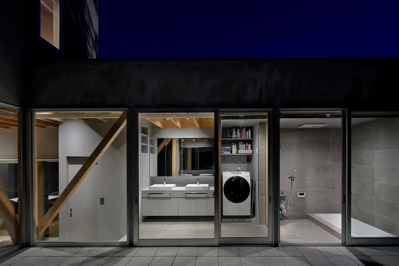 An evening view through sliding glass doors reveals a minimalist bathroom with a double vanity, a washing machine, and grey tile floors.
