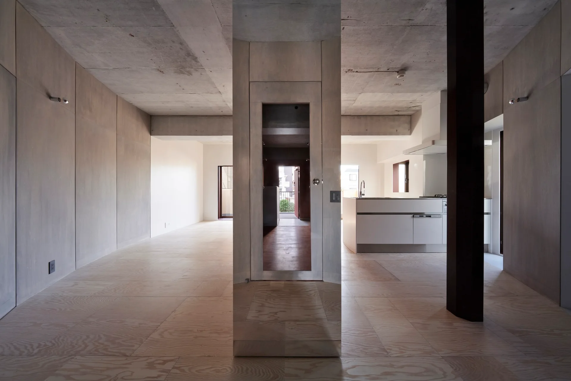 A minimalist apartment interior featuring light plywood walls and flooring, a raw concrete ceiling, a central mirrored column, and a white kitchen unit.