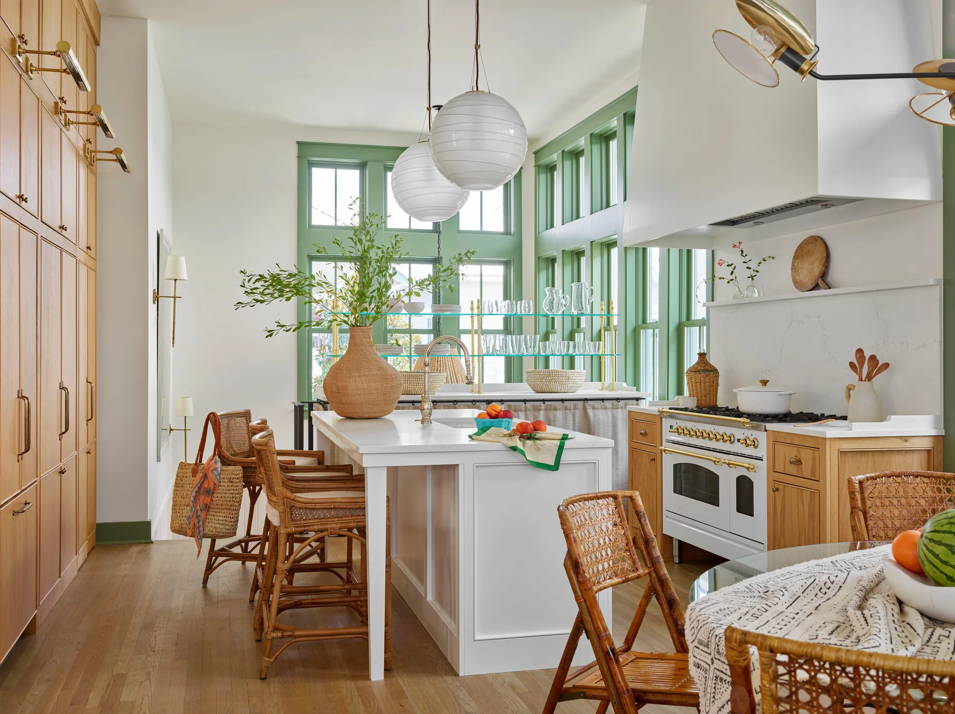 A bright kitchen features a white central island with rattan stools, light wood cabinets, and large green-trimmed windows.