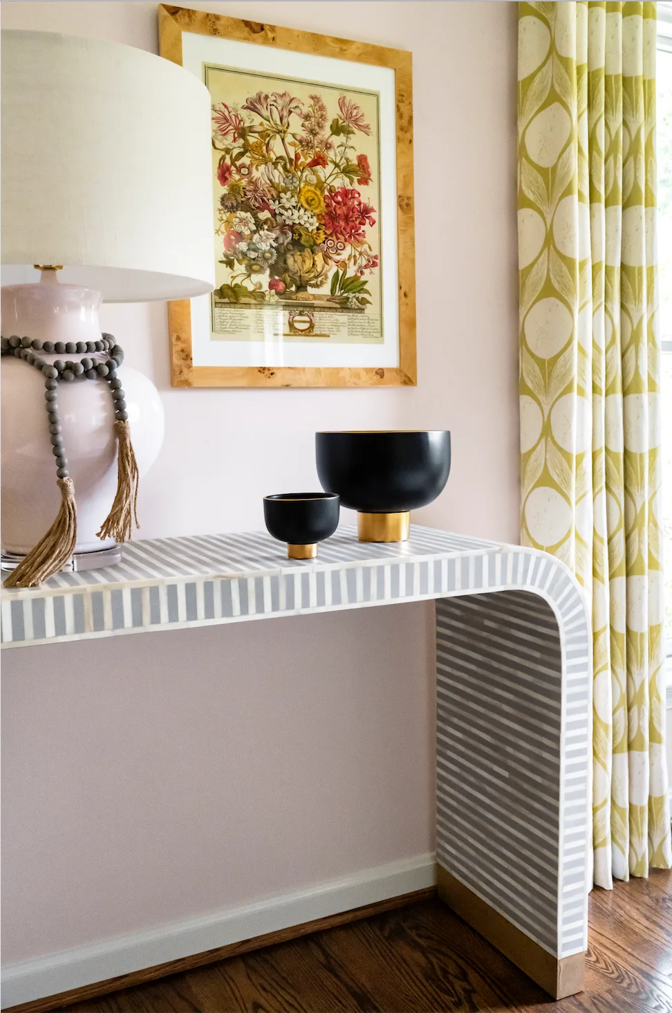 A striped waterfall console table sits against a pink wall next to patterned yellow drapes, displaying a pink lamp and two black decorative bowls.