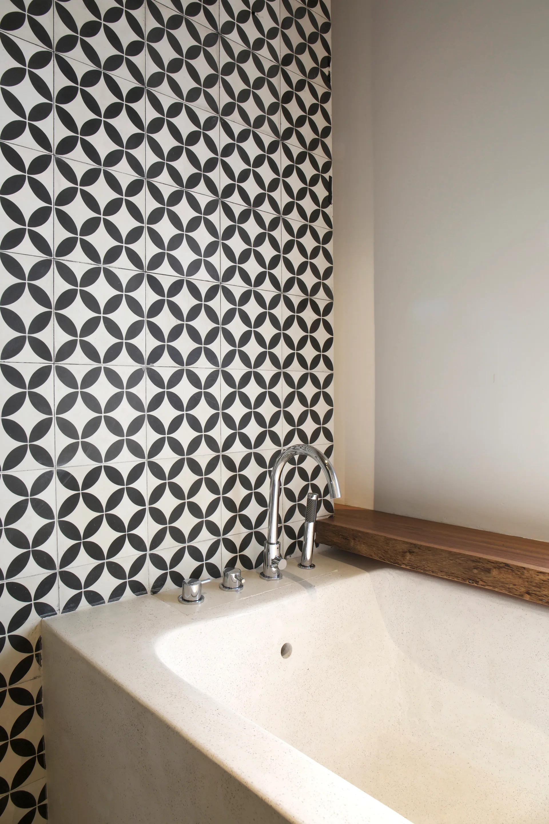 A built-in stone-like bathtub features a chrome arched faucet, set against a black and white geometric tiled wall and a natural wood shelf.