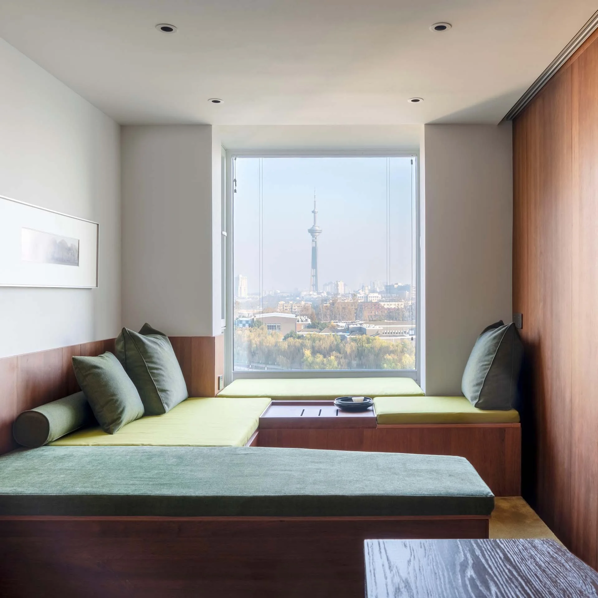 A custom wooden platform bed with green cushions and an integrated side table sits below a large window overlooking a city tower.