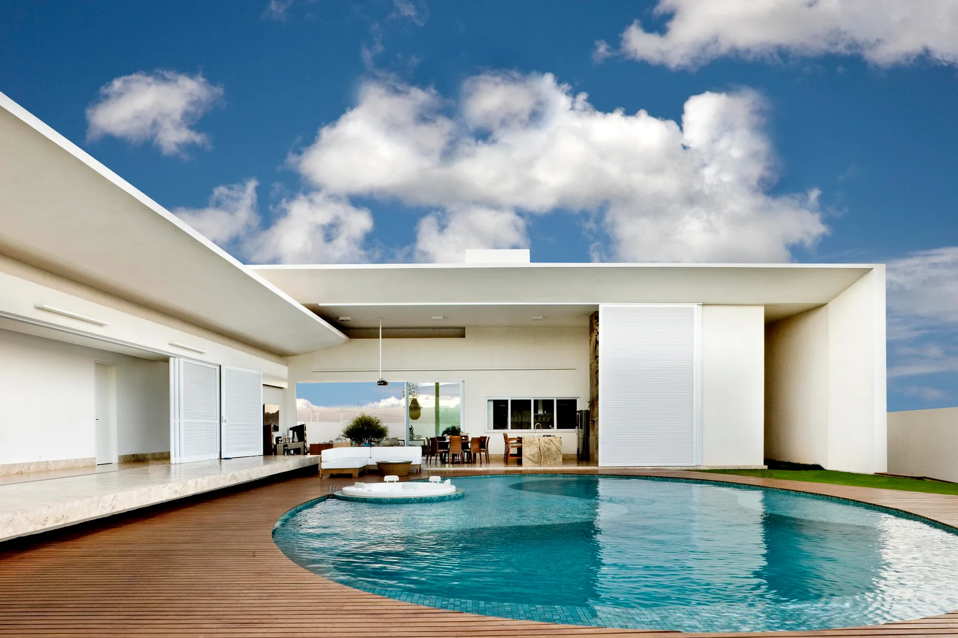 A modern white house features a curved swimming pool set into a wooden deck with an integrated spa and an open-air living pavilion.