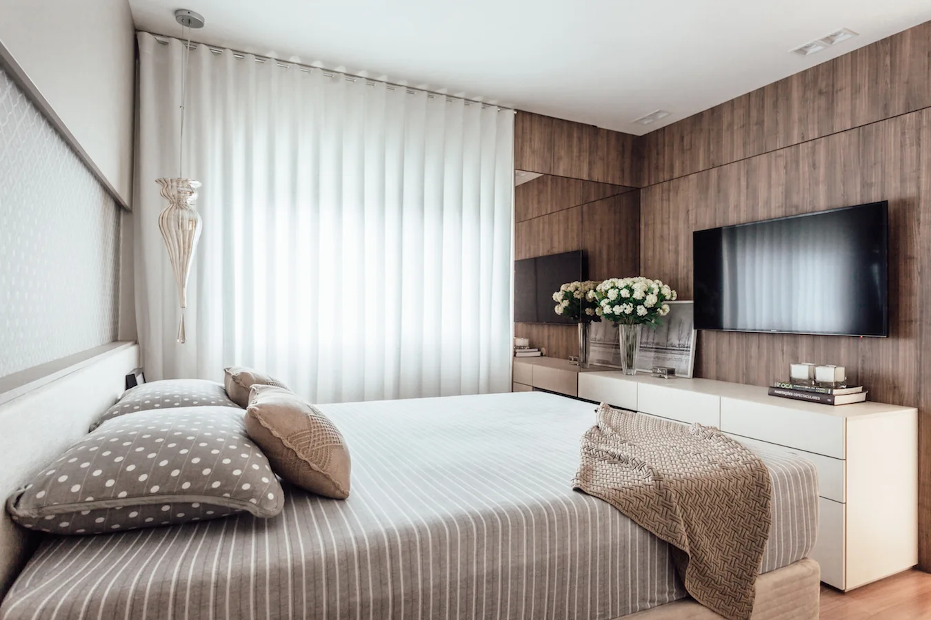 A modern bedroom features a bed with striped bedding next to a wood-paneled accent wall with a mounted television and a white console table.
