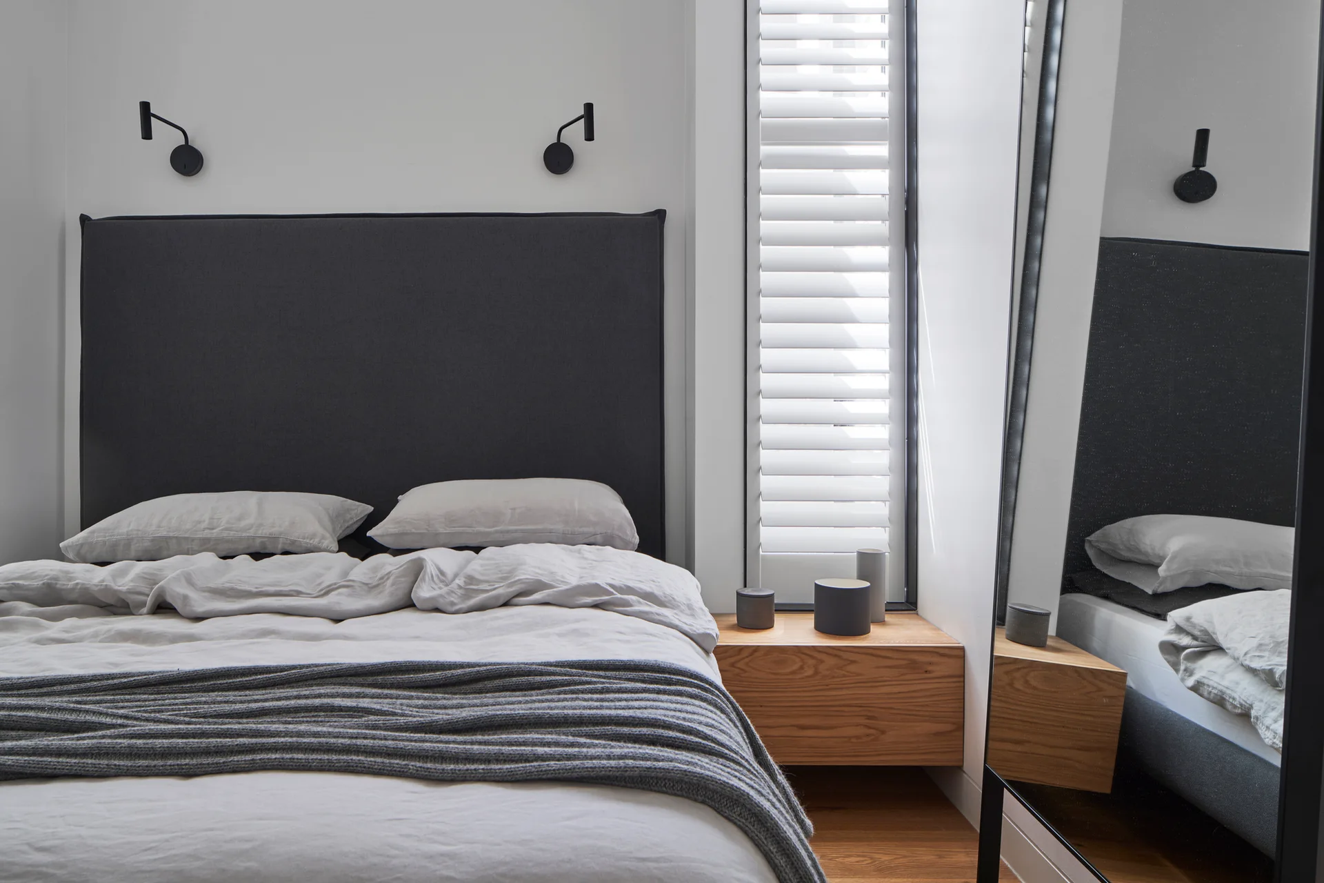 A modern bedroom features a dark grey upholstered headboard, rumpled light grey bedding, a floating wooden nightstand, and a tall floor mirror.