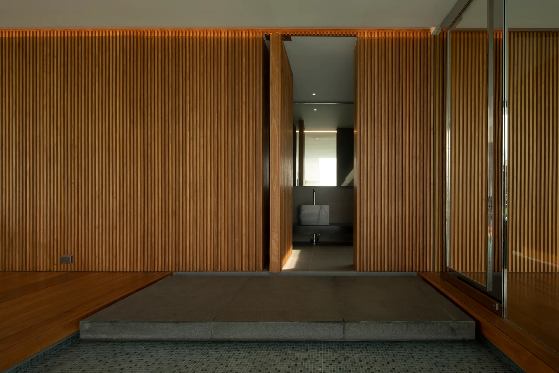 A vertical wood-slatted wall features a central pivot door opening to a bathroom, with a grey stone threshold step at the base.
