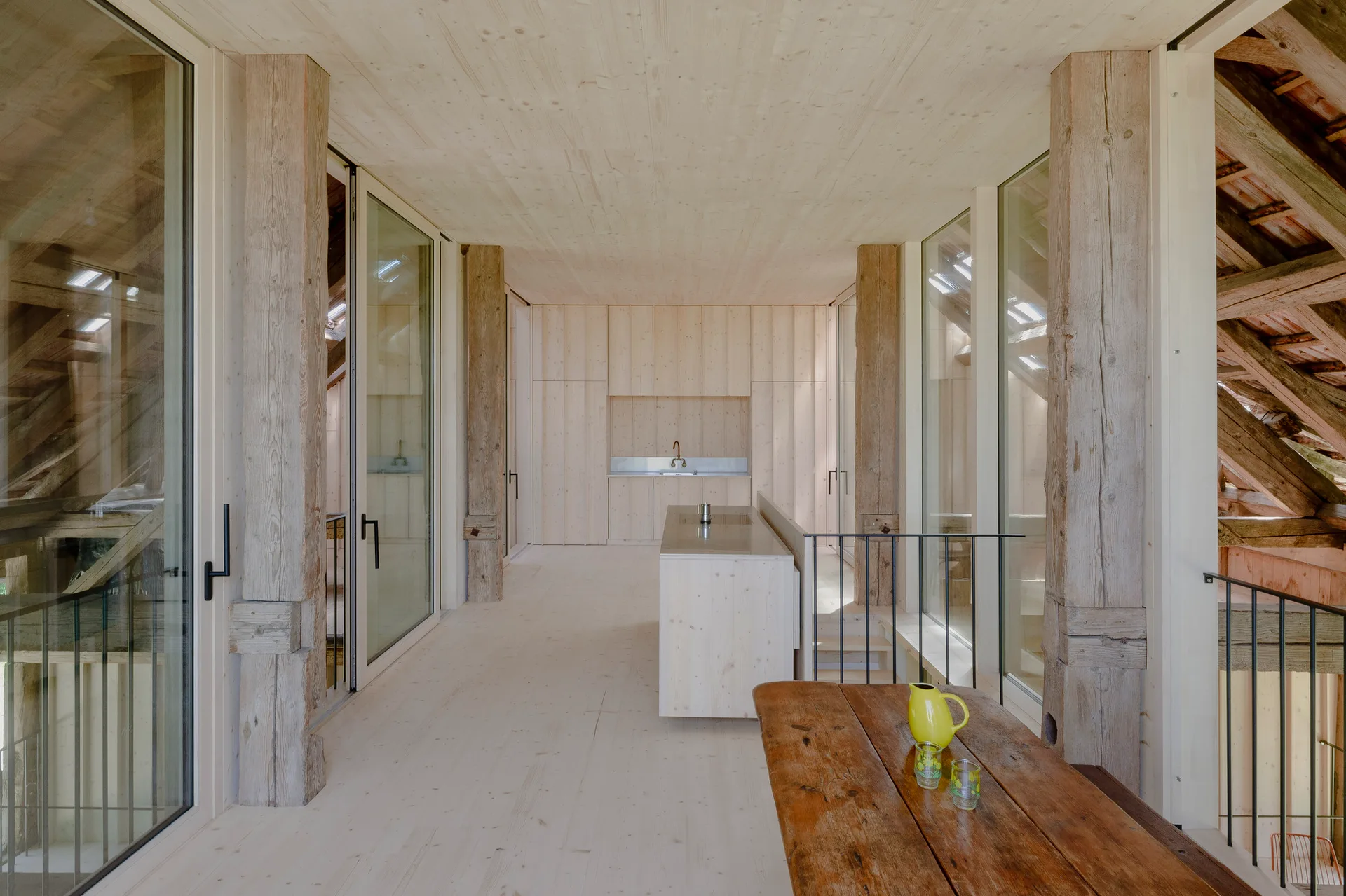 A minimalist kitchen features light wood paneled walls and ceilings, massive weathered timber support posts, and glass walls looking out to exposed rafters.