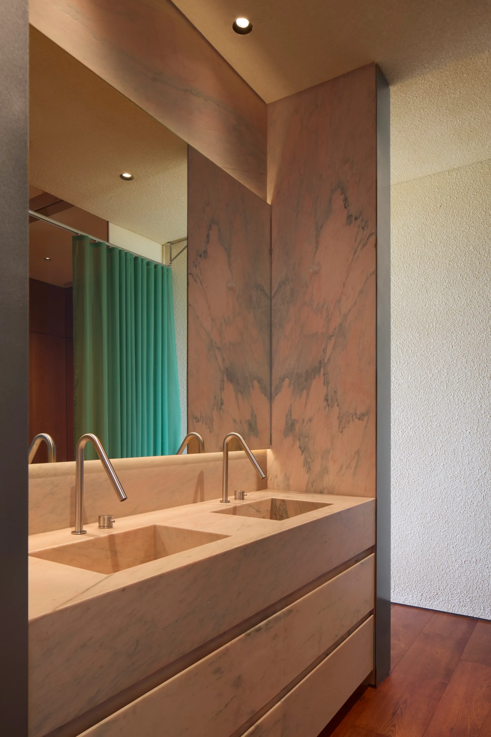 A modern pink marble bathroom vanity with two integrated sinks, curved silver faucets, and a large mirror reflecting a green curtain.