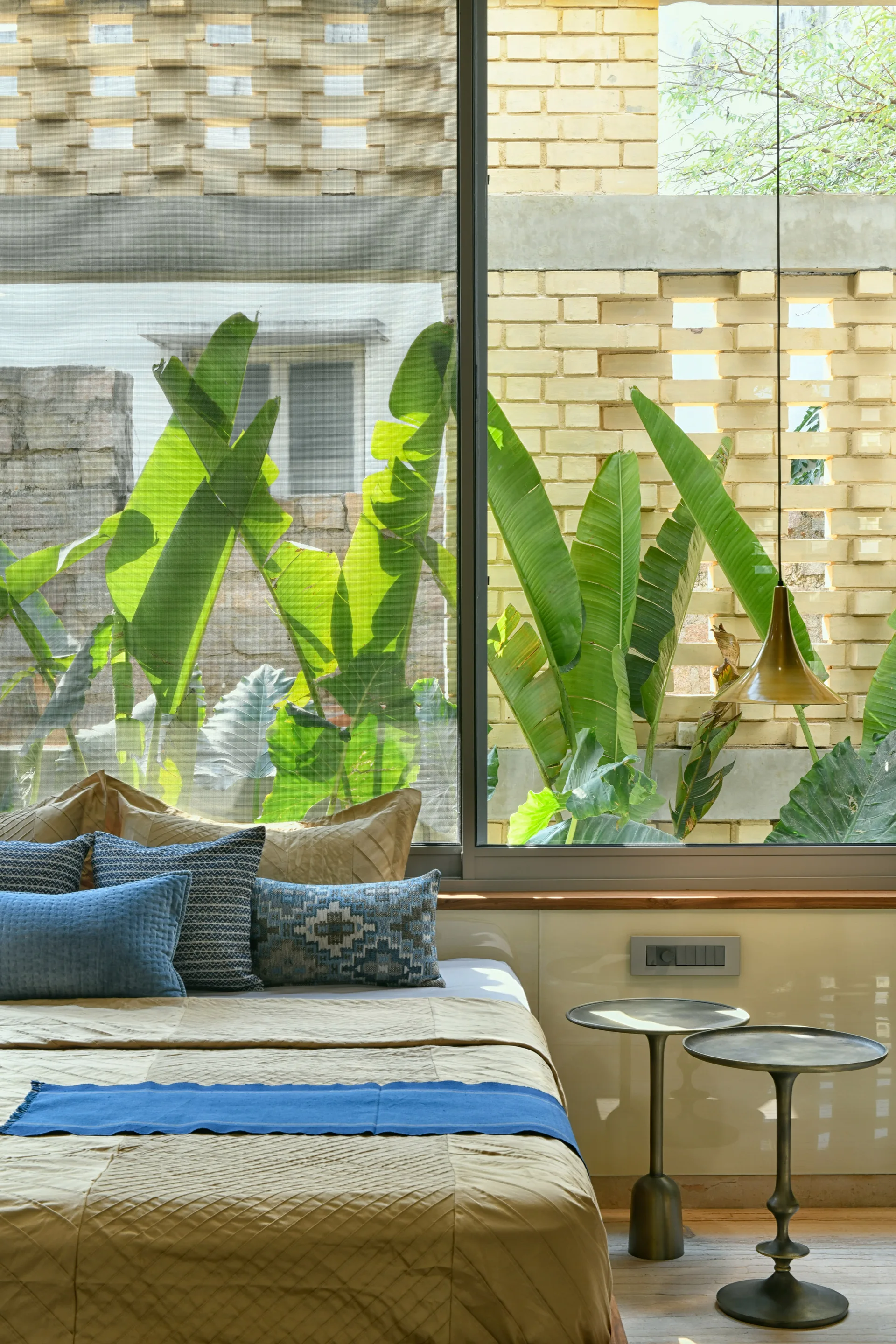 A bedroom features a bed with blue accents next to two pedestal side tables in front of a window showing a garden and brick screen.