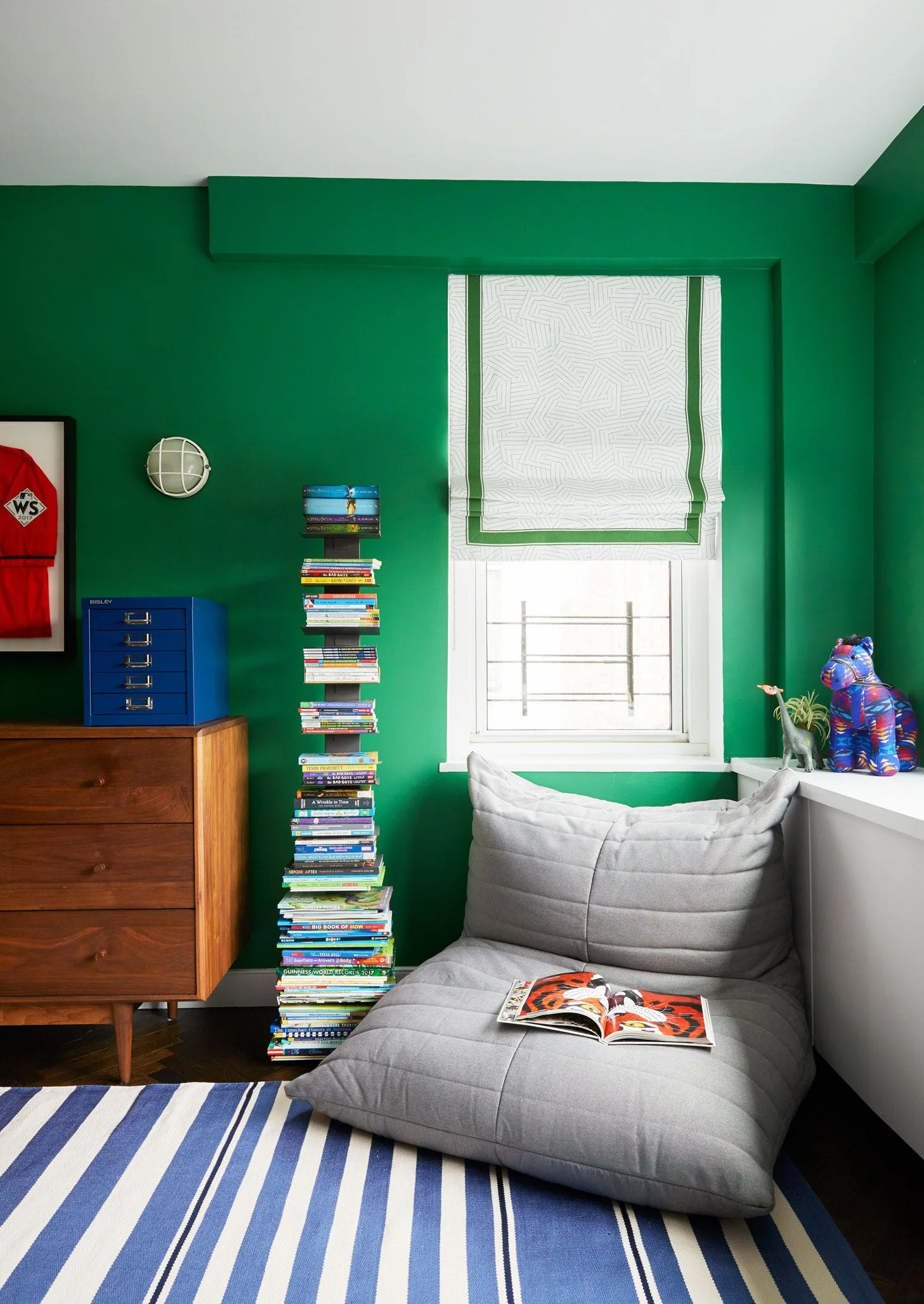 A kids' bedroom nook with vibrant green walls, a grey floor cushion, a vertical spine bookcase, a wooden dresser, and a blue and white striped rug.