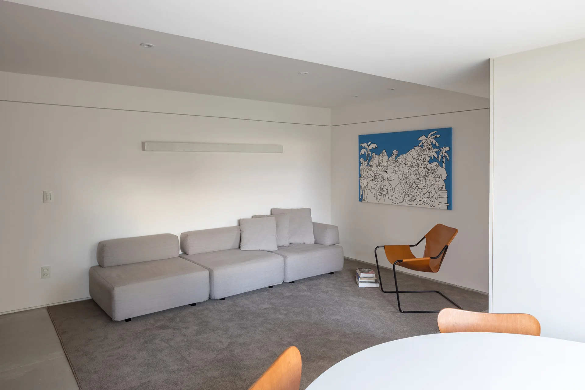 A minimalist living room featuring a low light gray modular sofa, a tan leather armchair on a gray carpet, and a large blue painting on a white wall.