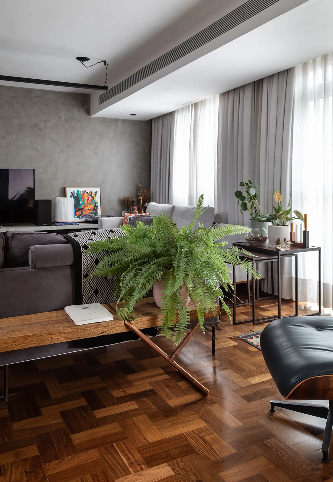 A modern living room features a grey sofa, herringbone parquet wood flooring, a grey plaster wall, and a wooden bench holding a large green fern.