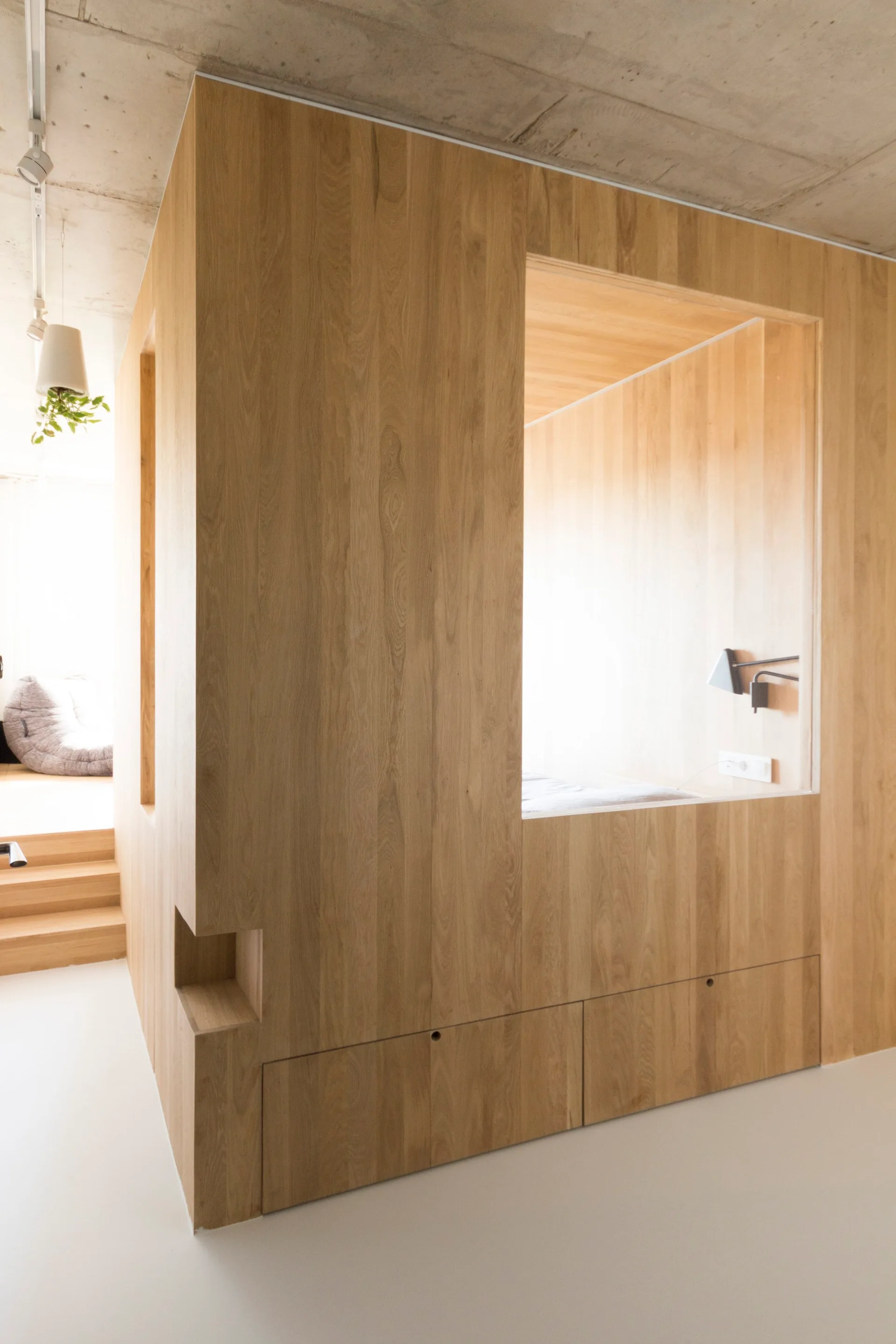 A minimalist wooden partition structure featuring a built-in sleeping nook, storage drawers at the base, and an exposed concrete ceiling above.