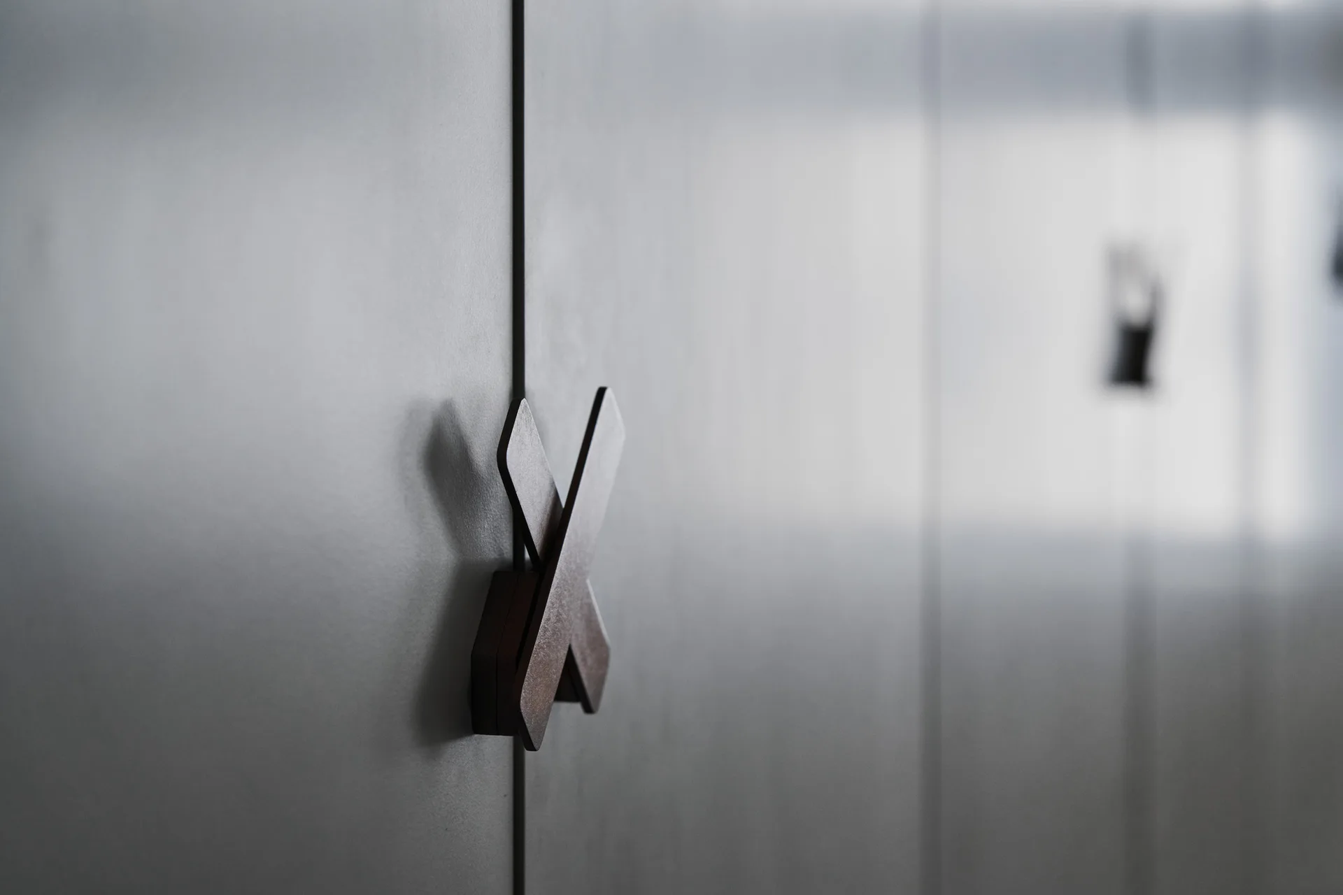 A close-up of a unique dark wood X-shaped handle mounted on a flat matte grey cabinet door.