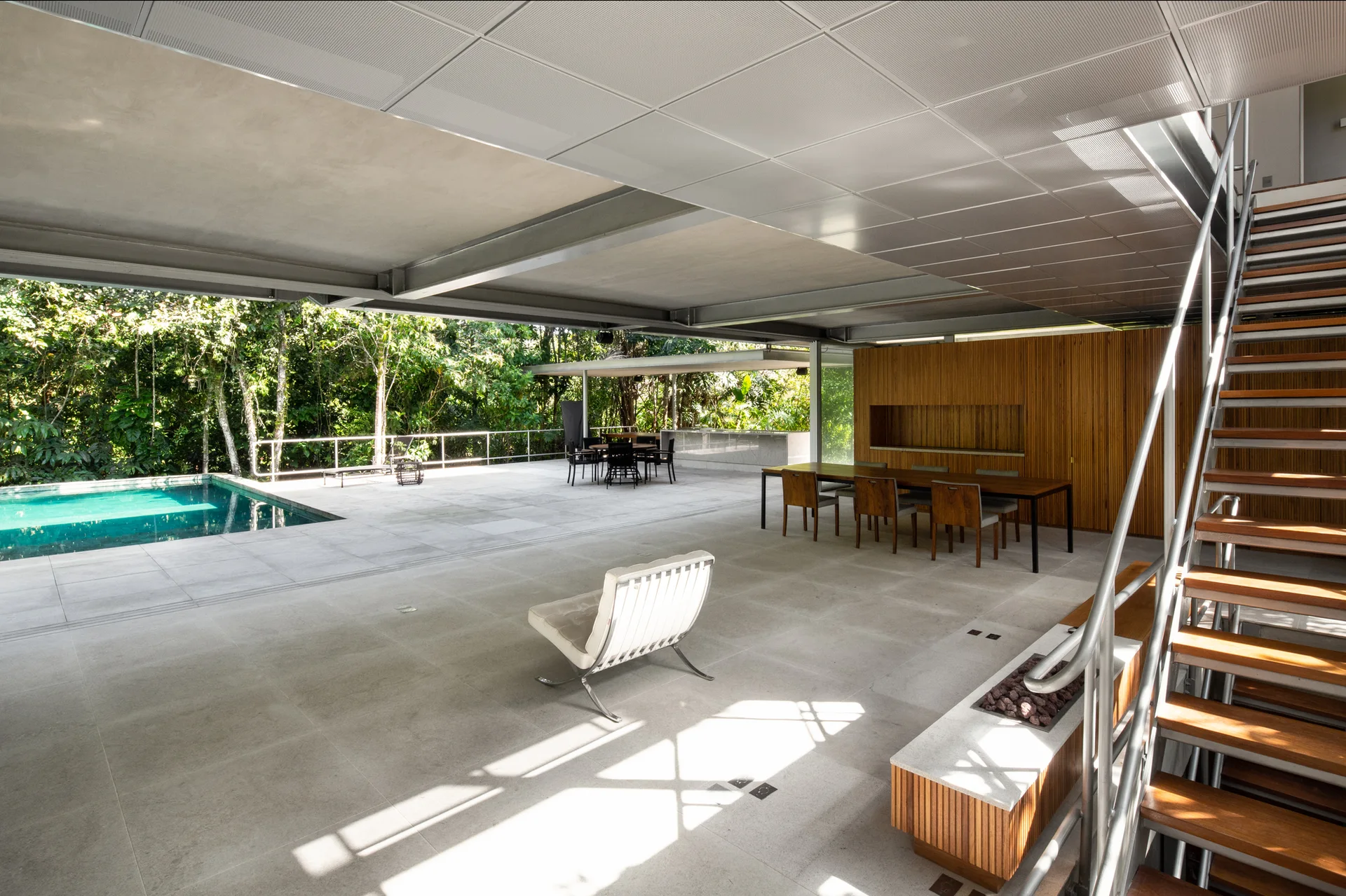A spacious modern open-air patio features a rectangular swimming pool, concrete tile flooring, an open-riser staircase, and a slatted wood wall.