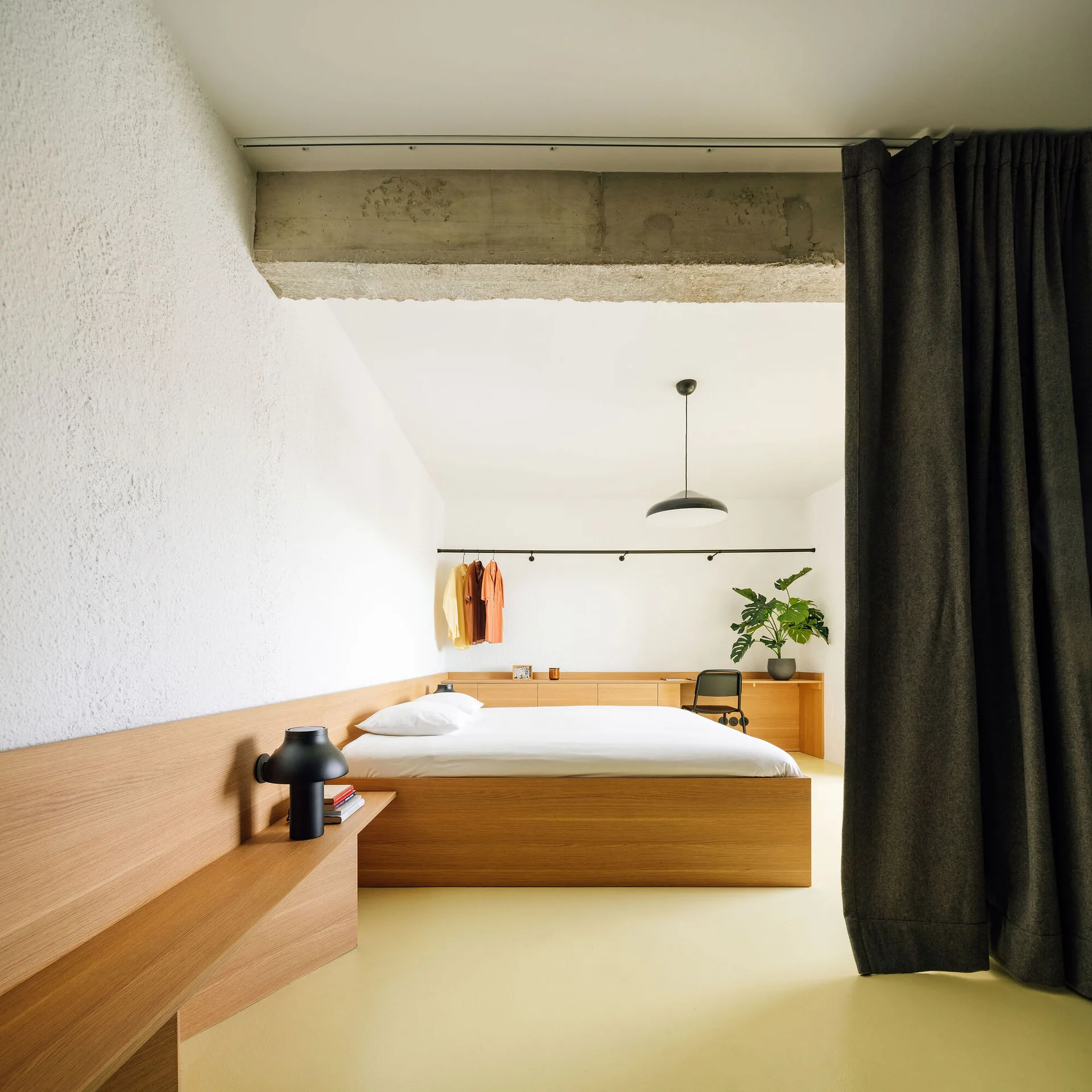A minimalist bedroom features a low wooden platform bed integrated with a matching wall-wrapping shelf and built-in desk under an exposed concrete beam.