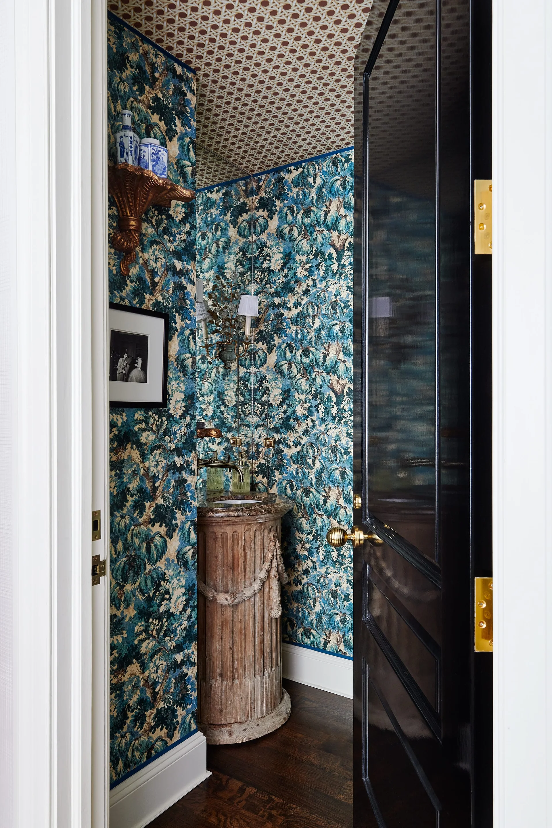 A small powder room features dense botanical wallpaper, a carved wood pedestal sink, a geometric patterned ceiling, and a high-gloss black door.