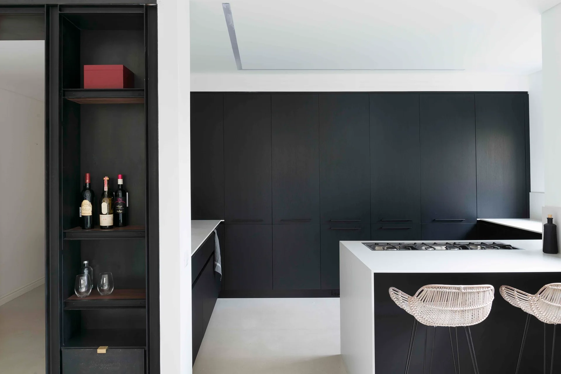 A minimalist kitchen features matte black floor-to-ceiling cabinets, a white island with a built-in cooktop, and two woven bar stools.