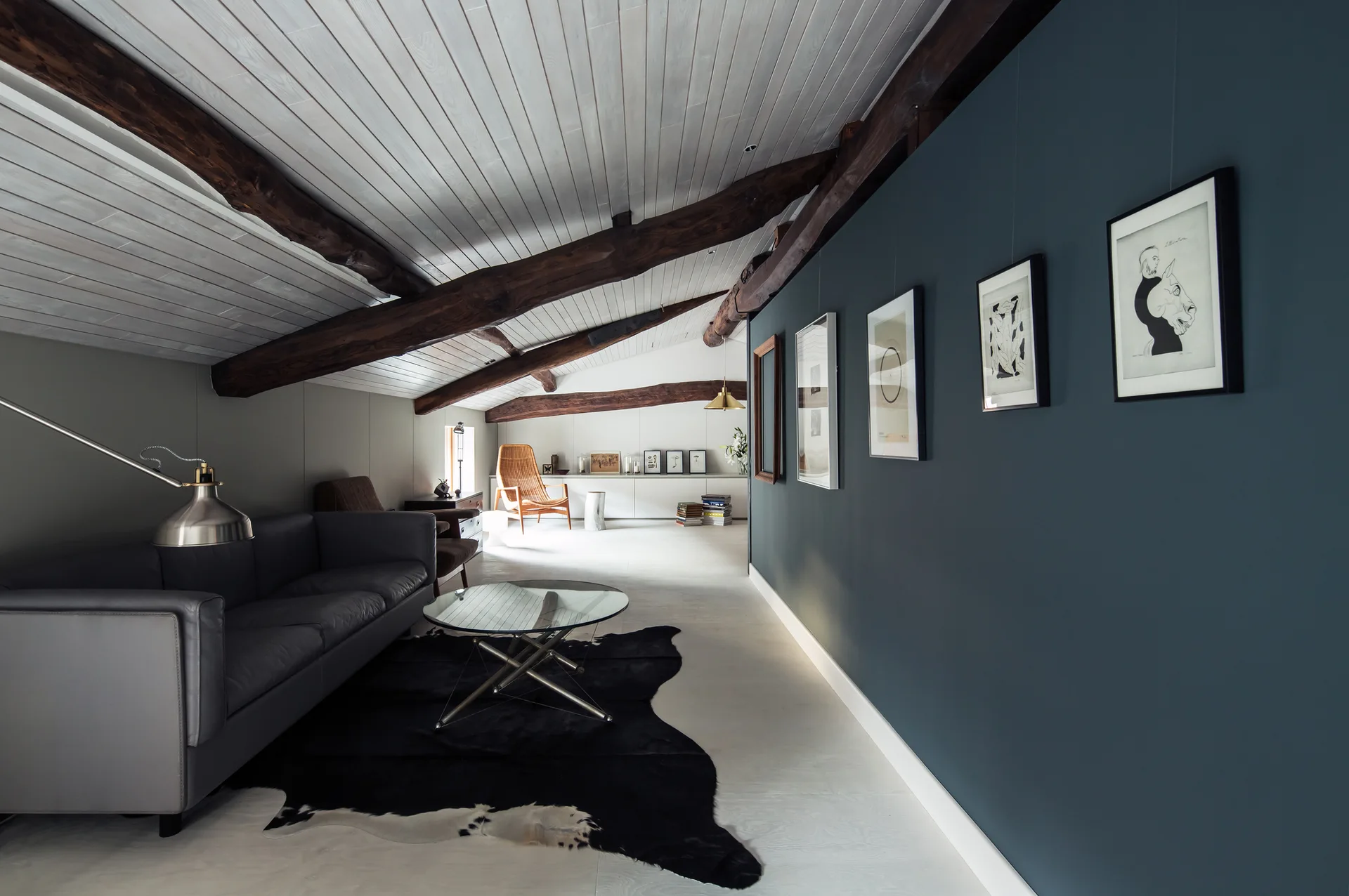 An attic living room featuring a grey leather sofa on a cowhide rug, rustic wood ceiling beams, and a deep teal wall with framed art.