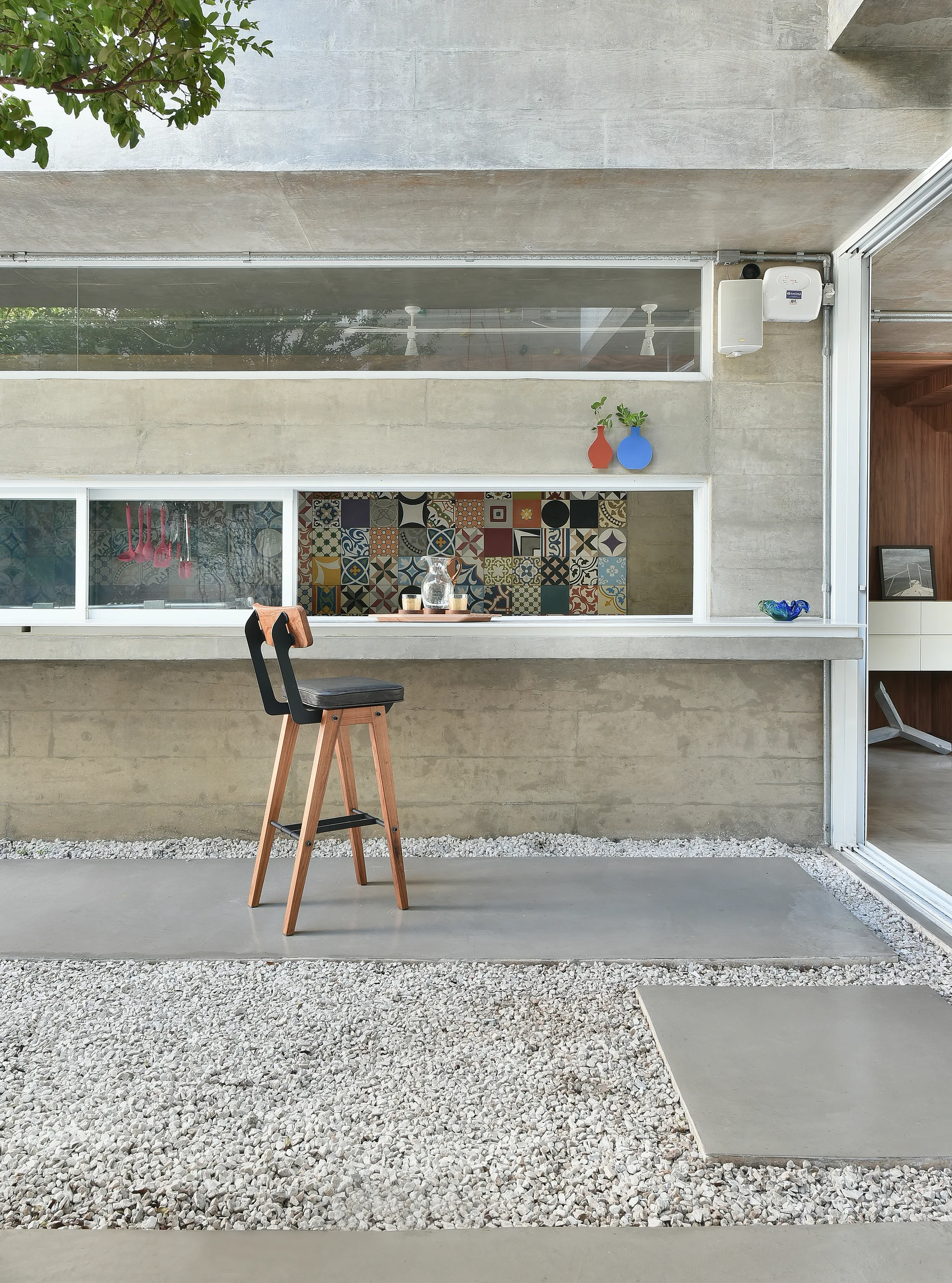 A wooden bar stool sits by a concrete outdoor counter with a pass-through window looking into a kitchen with a colorful mosaic backsplash.