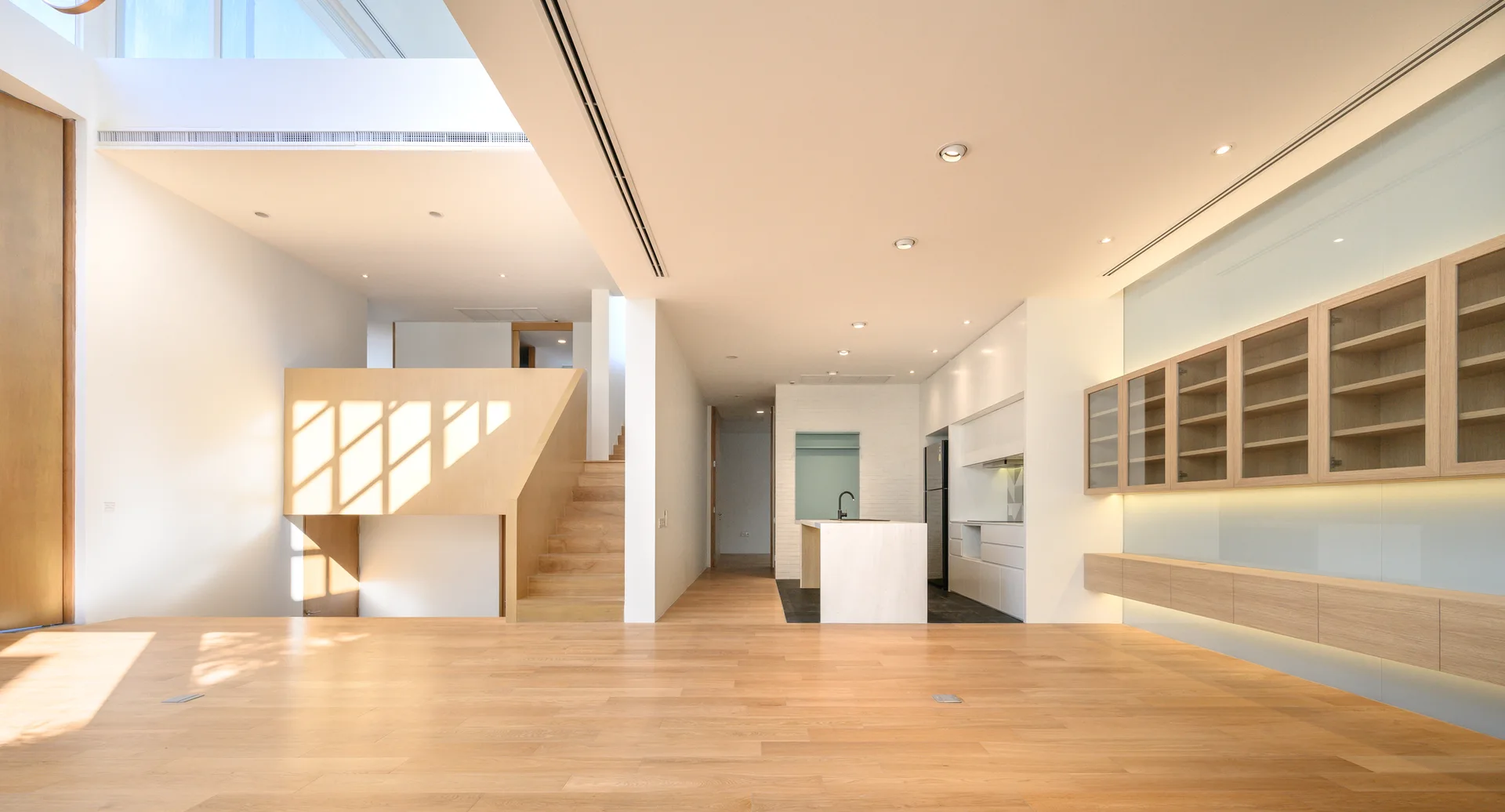 A spacious, open-concept living area with light wood floors, white walls, a wooden staircase, and built-in glass-front cabinets.