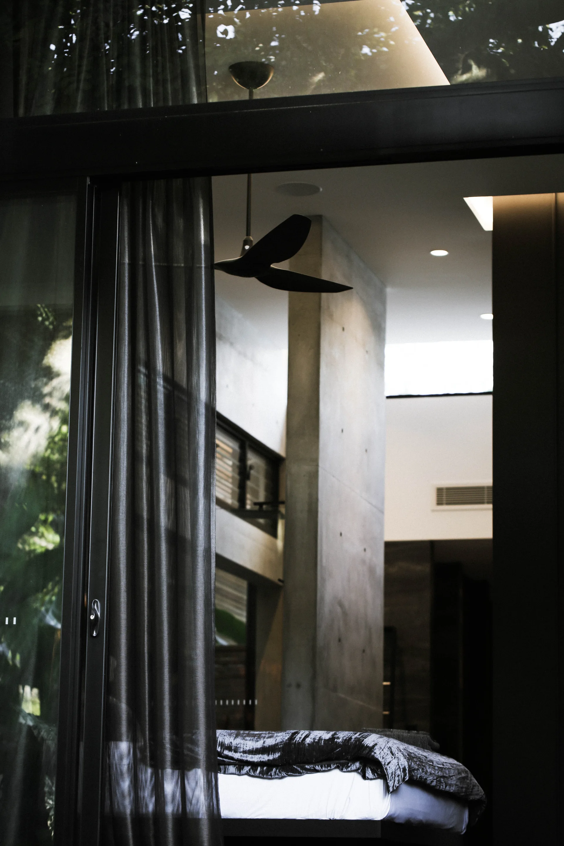 A modern bedroom viewed through glass sliding doors with sheer dark curtains featuring a bed and a large concrete column.