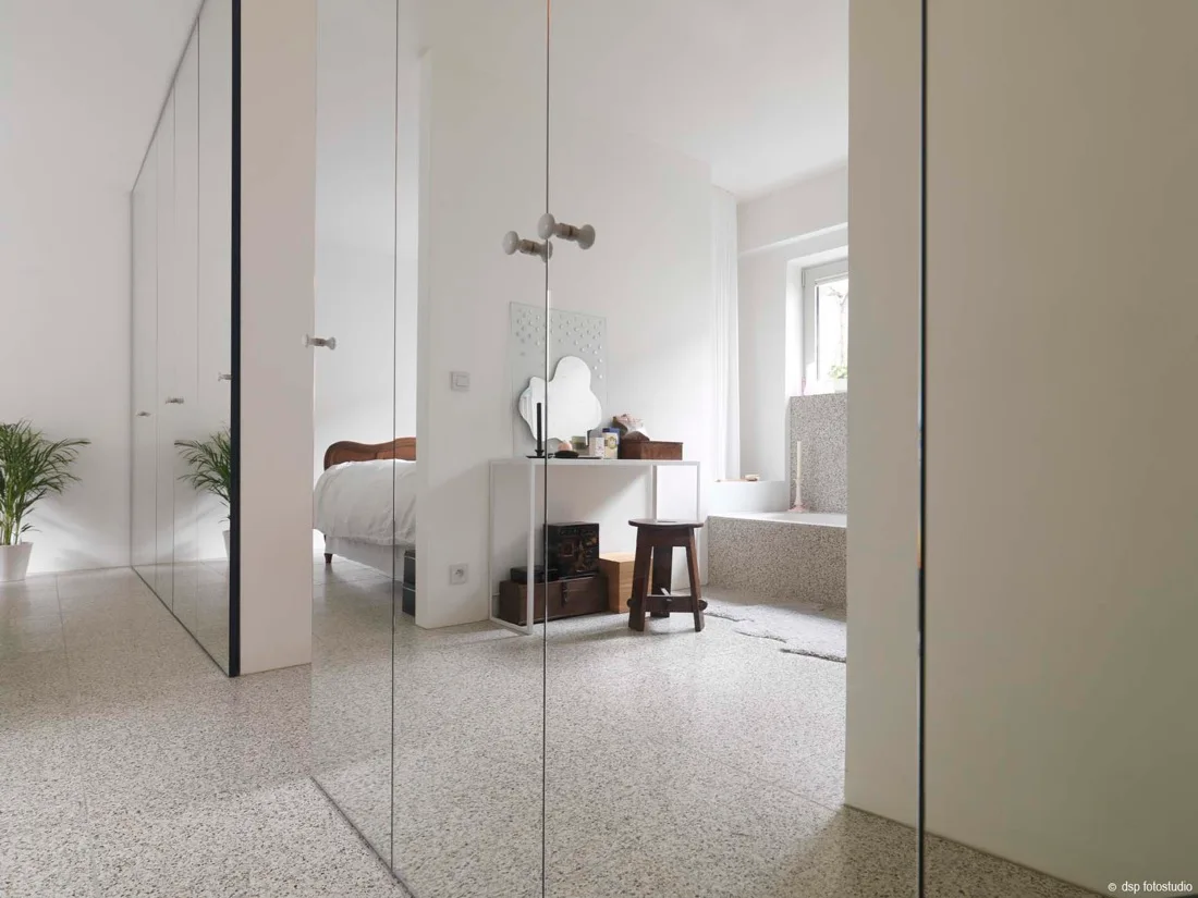 A minimalist white bedroom suite features flecked grey terrazzo flooring, a built-in bathtub, and full-height mirrored closet doors.