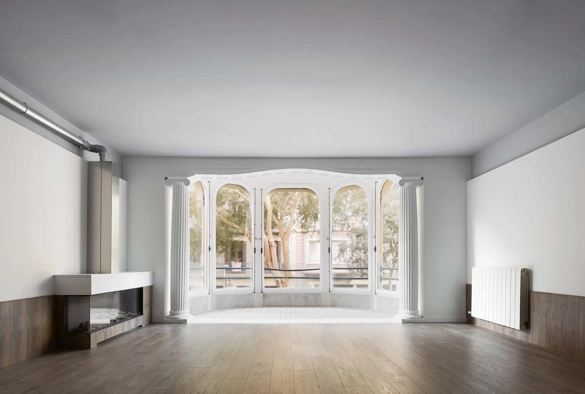An empty room featuring a large arched bay window flanked by white fluted columns, wood plank flooring, and a modern metal fireplace.
