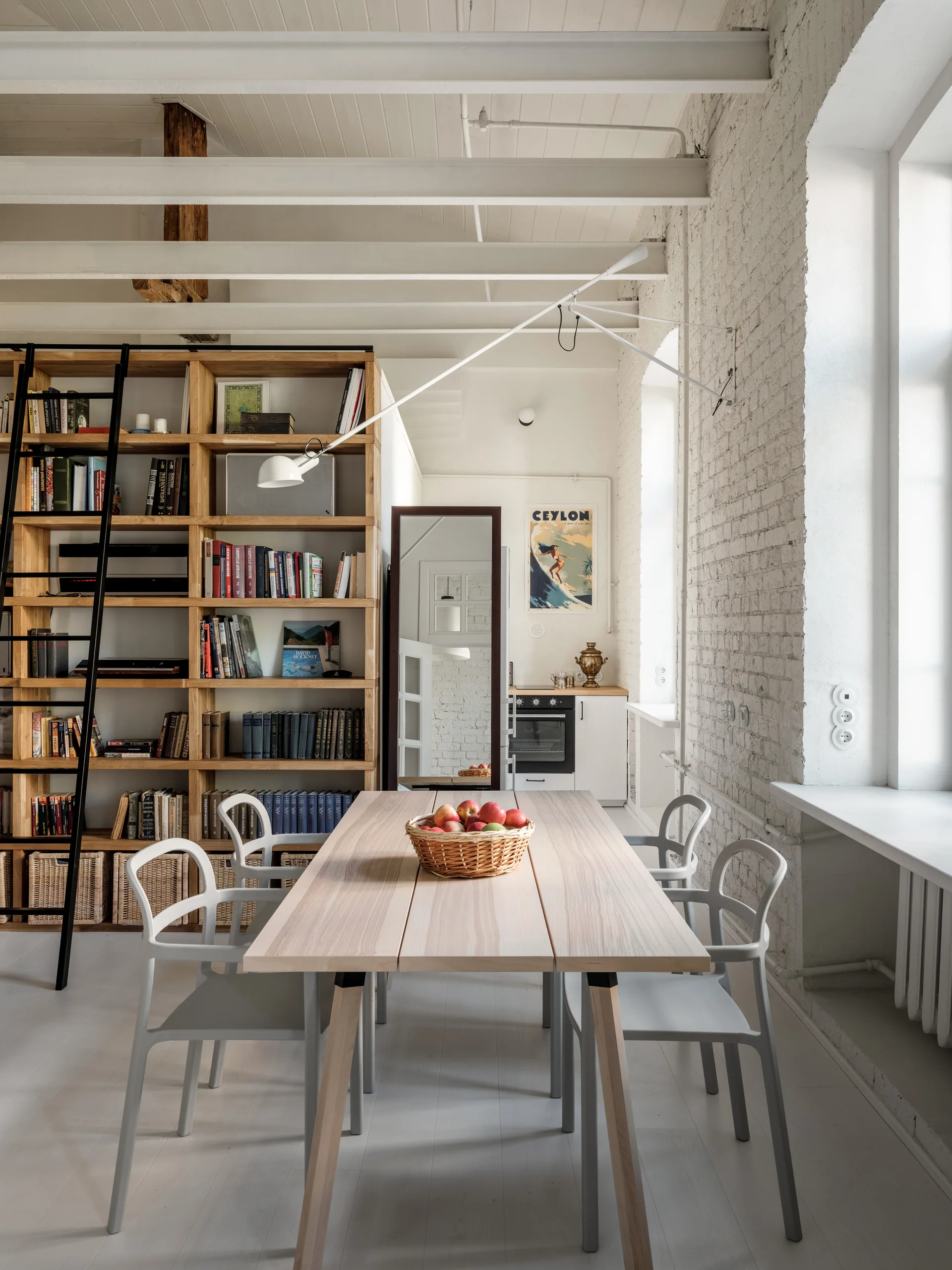 A light wood dining table with chairs sits in a loft dining area beside a white painted brick wall and a tall wooden bookshelf with a black ladder.