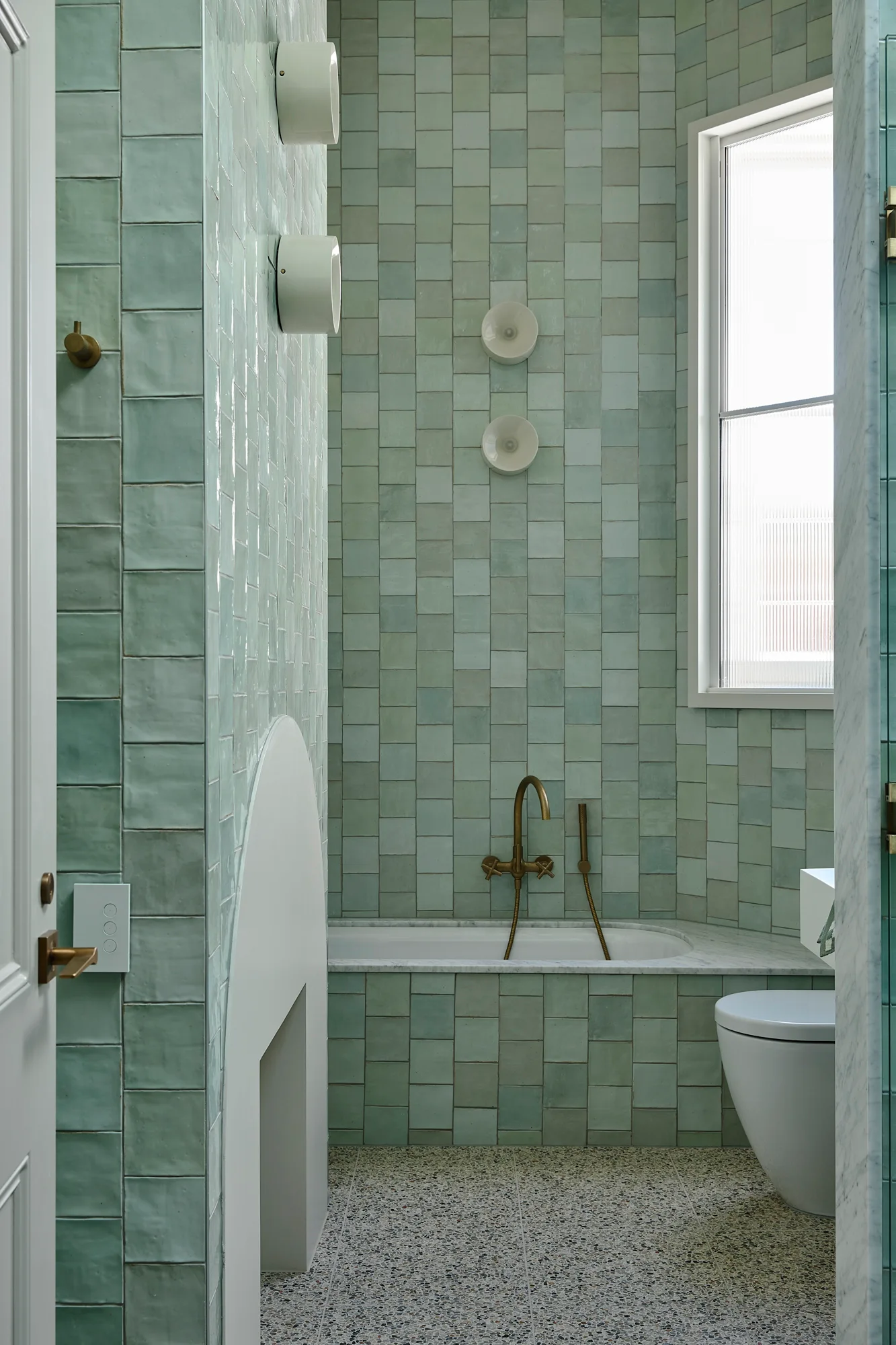 A contemporary bathroom features mint green square wall tiles, a built-in bathtub with brass fixtures, a modern toilet, and terrazzo flooring.